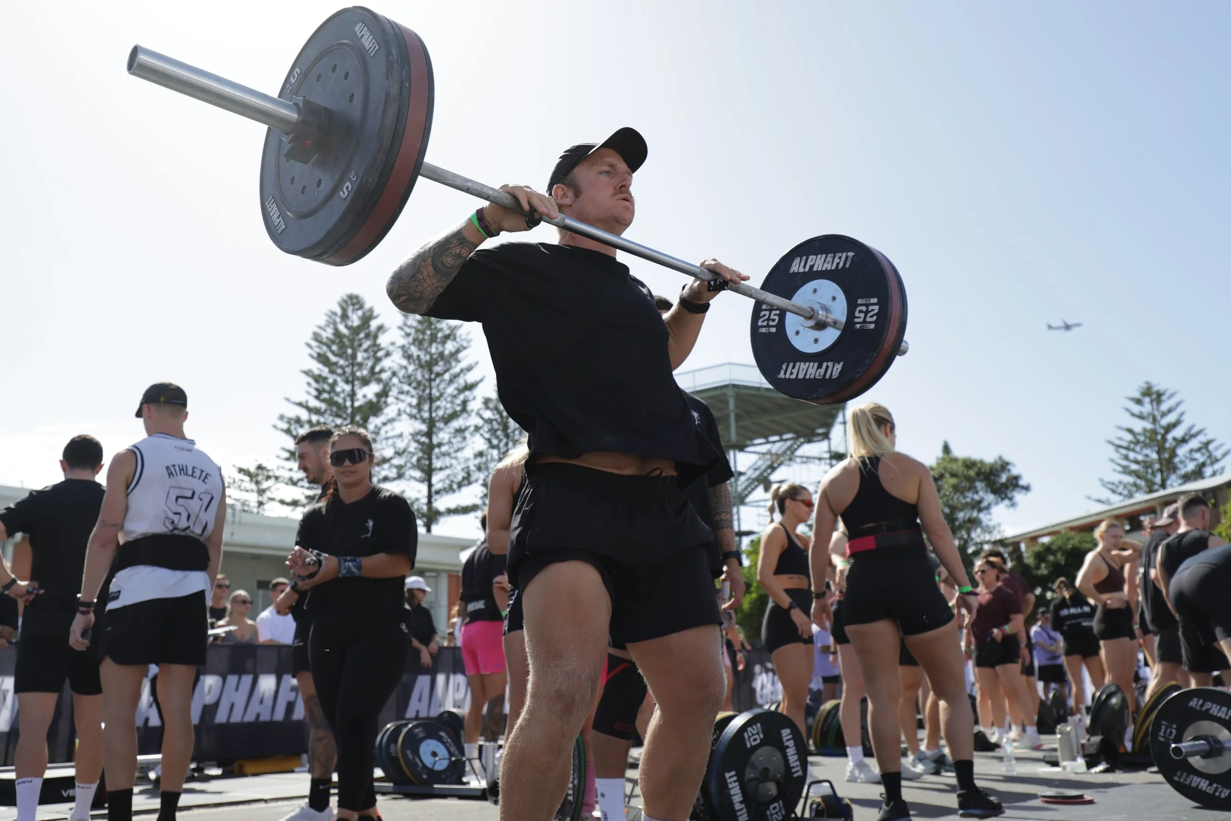 A man lifting a barbell at an outdoor fitness event with other participants and spectators around.