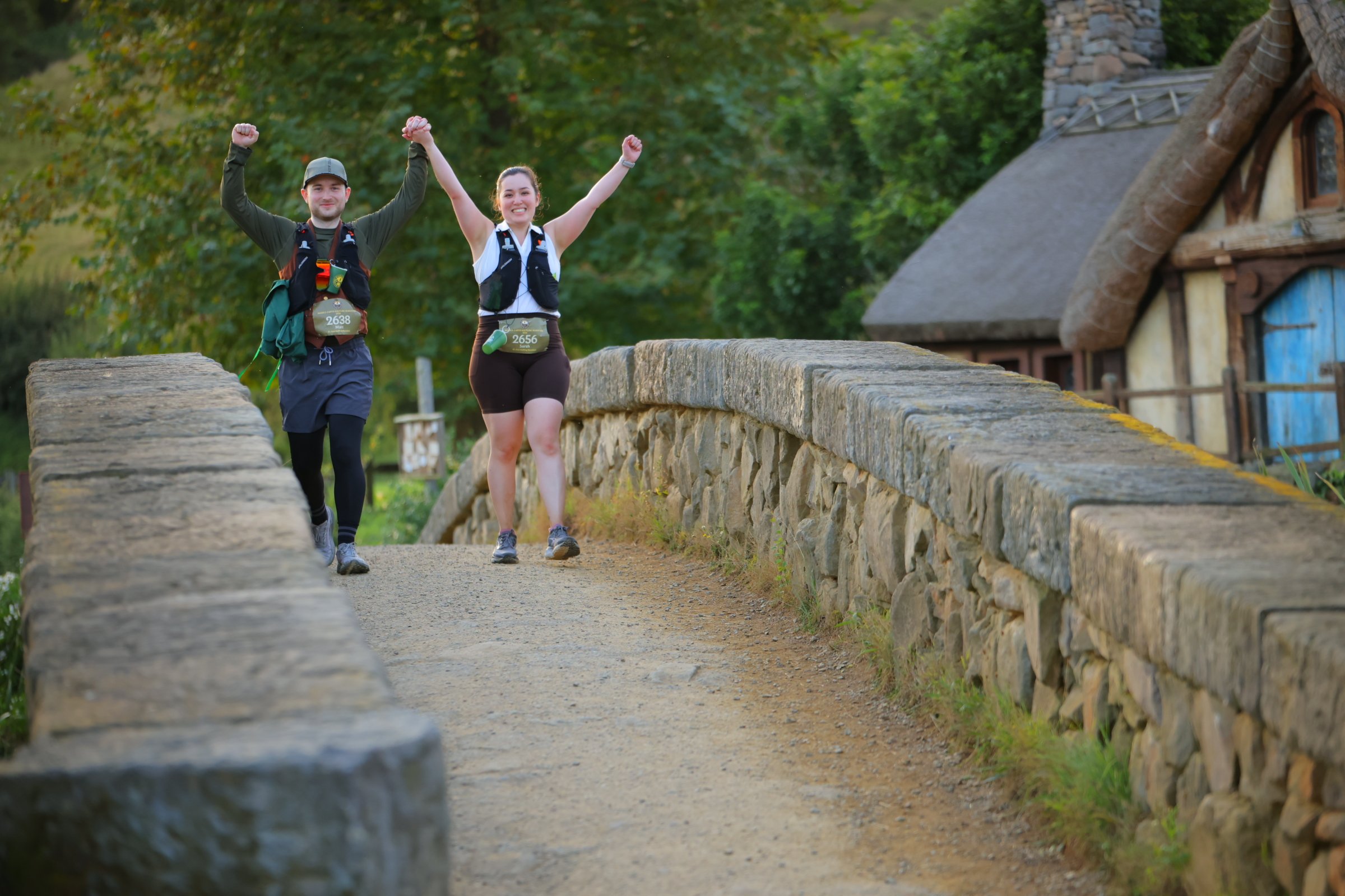 A man and woman celebrating crossing a finish line in a park or trail setting on a stone bridge, wearing running gear and race bibs, holding hands with their arms raised.