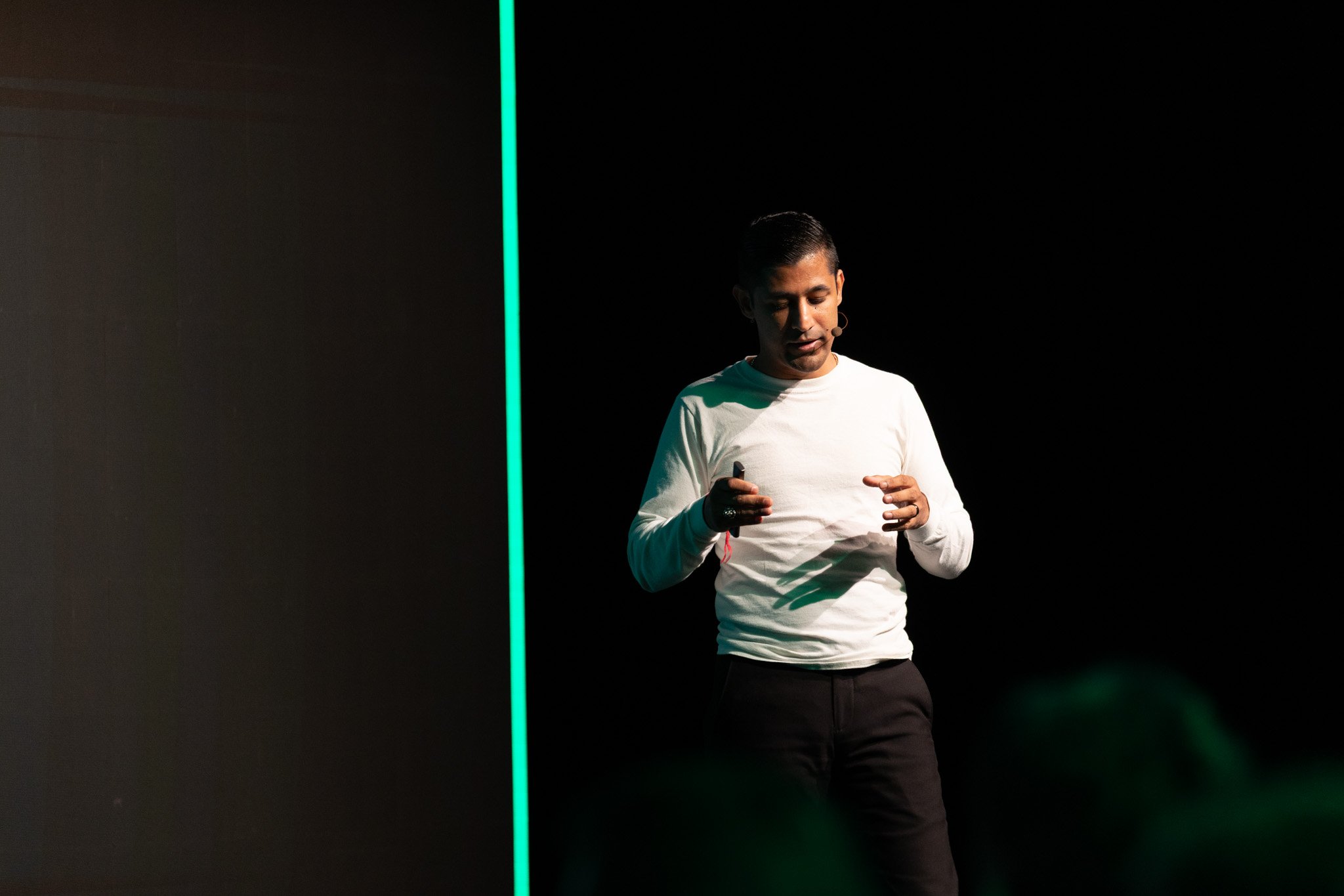 A man in a white shirt with a microphone headset looking down at his hands, standing against a dark background with a green vertical light line.