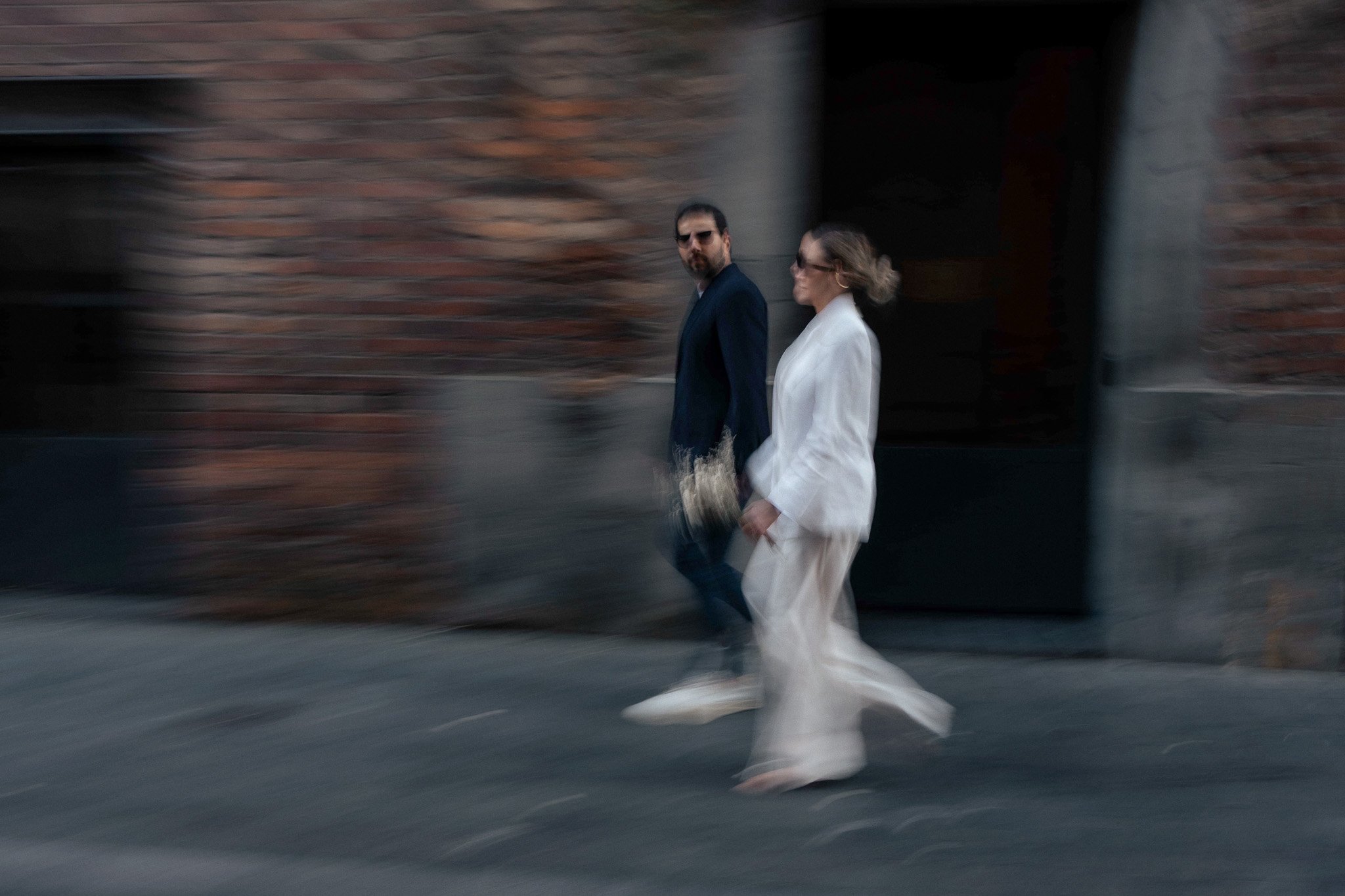 A man and woman walk together on a city sidewalk, blurred in motion, with a brick building in the background. The man wears dark sunglasses and a dark suit, and the woman wears a light-colored coat and sunglasses.