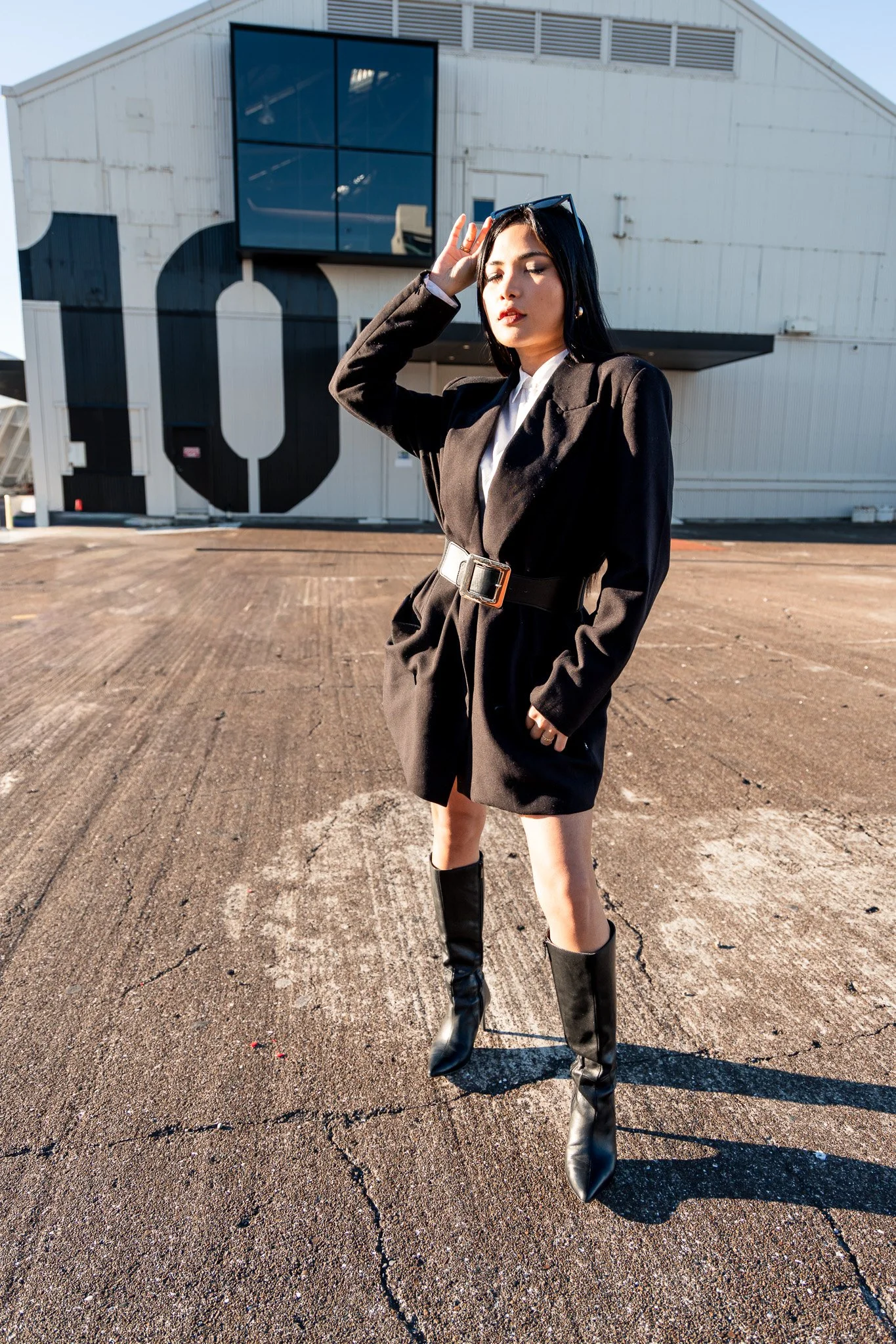 A woman with black hair dressed in a black blazer, white blouse, and black leather knee-high boots standing outdoors on a parking lot with a building in the background that has a large black number 10 painted on it.
