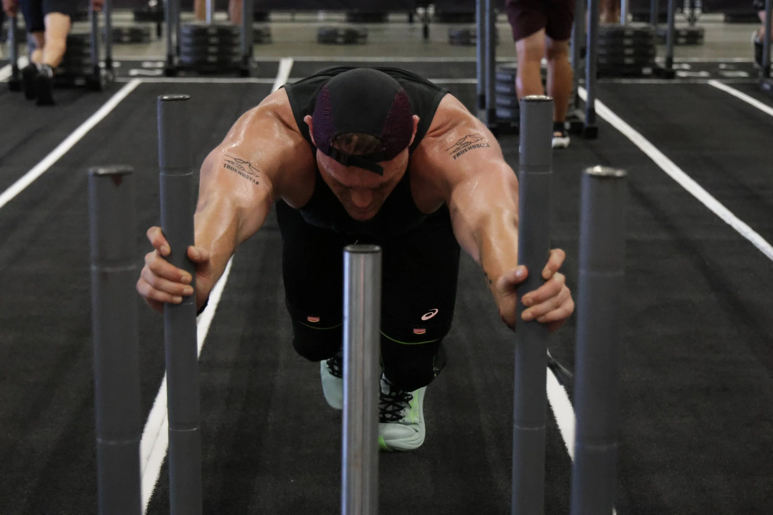 A man performing a push-up in a gym, wearing a black sleeveless shirt, a cap backwards, and neon green shoes, sweat visible on his skin.
