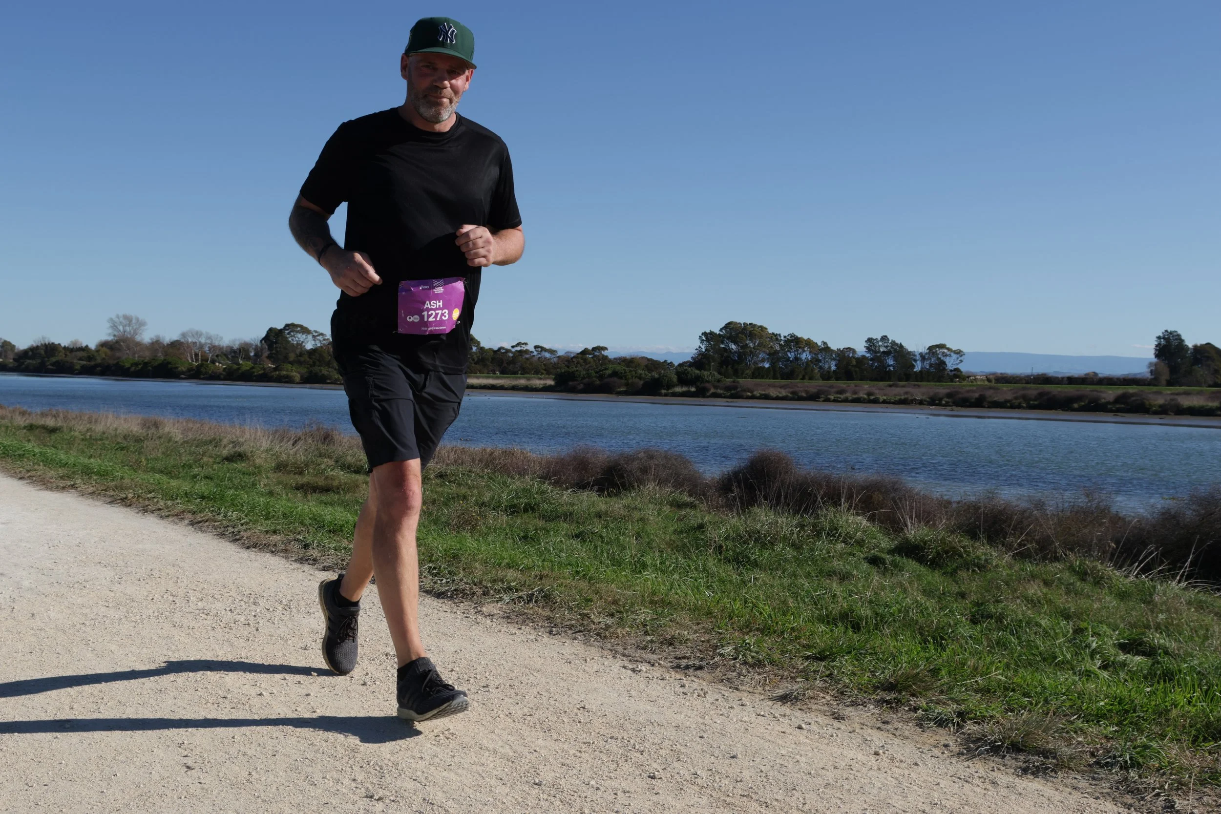 Man jogging along a dirt path next to a body of water, with trees and mountains in the background under a clear blue sky.