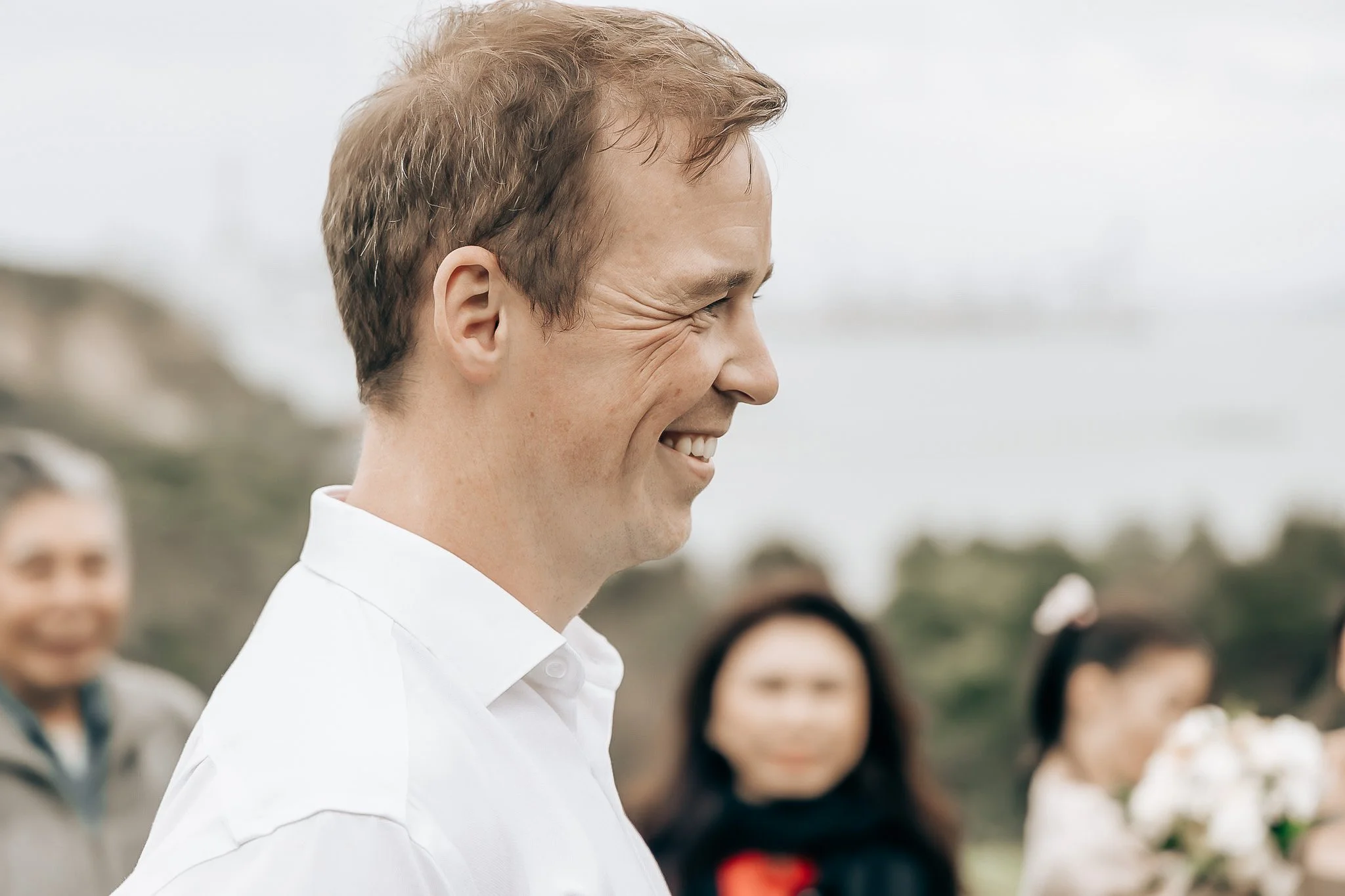 A man with light brown hair smiling, wearing a white shirt, outdoor setting with a group of people in the background.