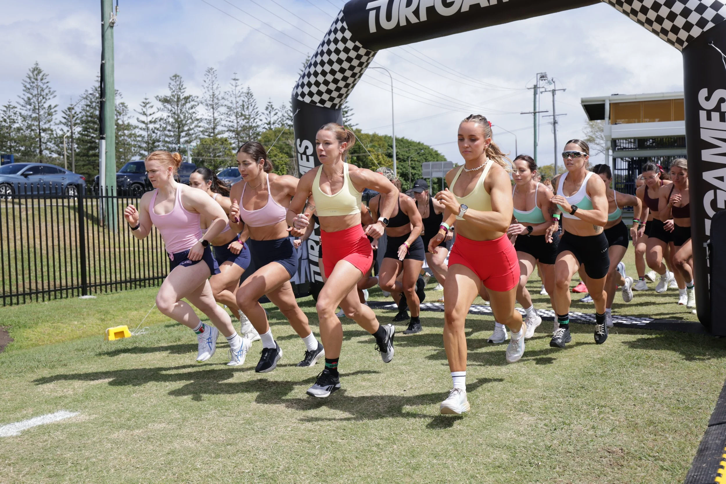 Women participating in a running race outdoors, starting from a marked line, under a checkered archway with 'TORREGA' written on it, with trees and parked cars in the background.
