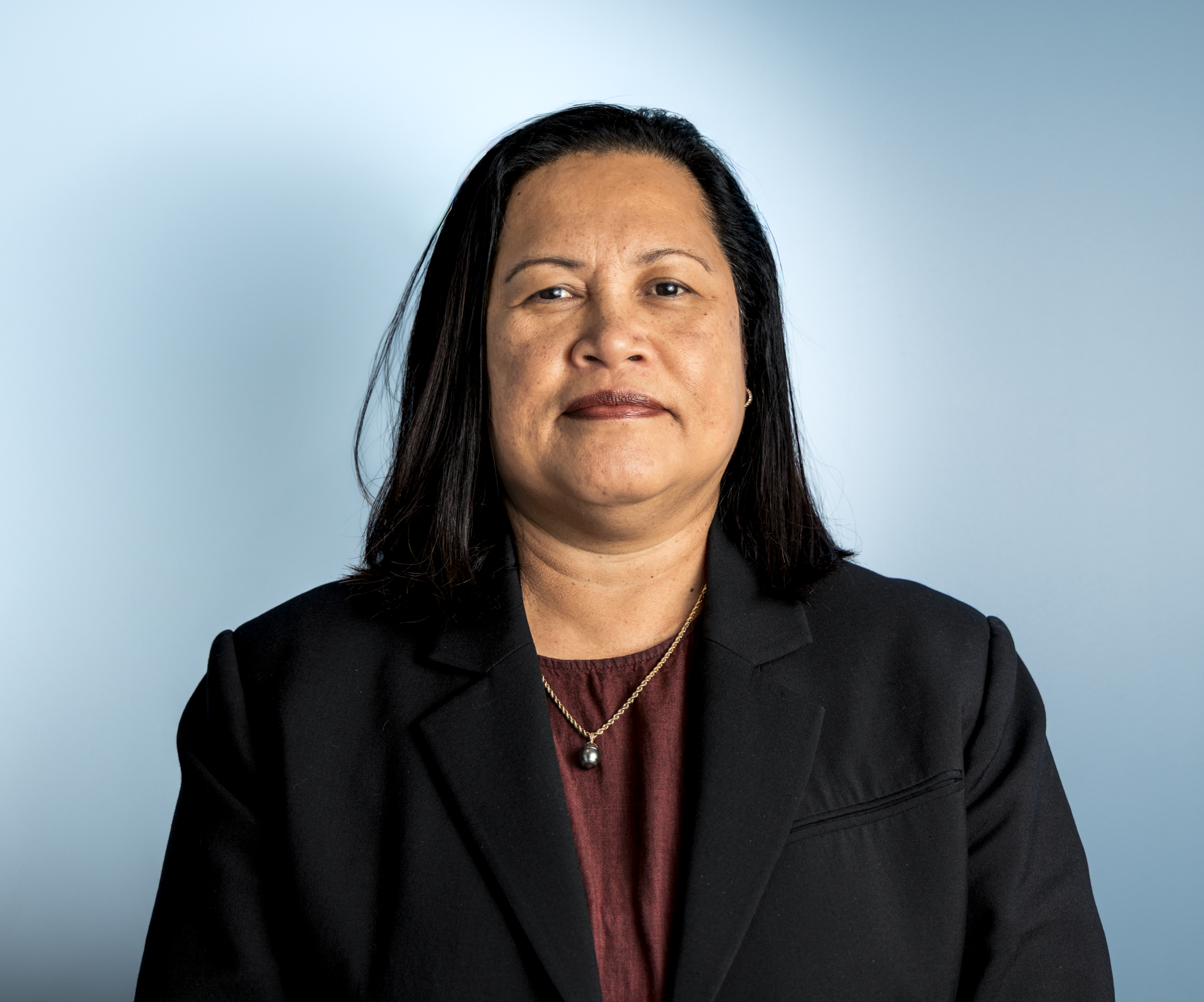 Close-up of a woman with black hair, wearing a black blazer and a red blouse, set against a light blue background.