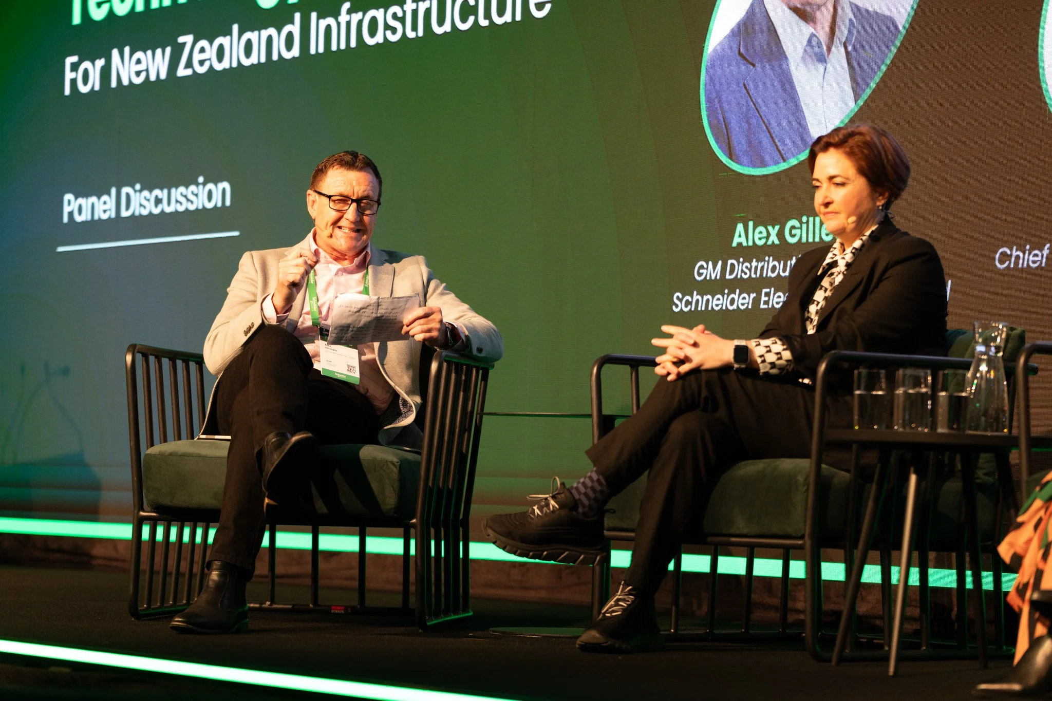 Two individuals, a man with glasses and a woman with short hair, sitting on a stage during a panel discussion at a conference. The background displays a green screen with the text "Technology for New Zealand Infrastructure" and names of the speakers.