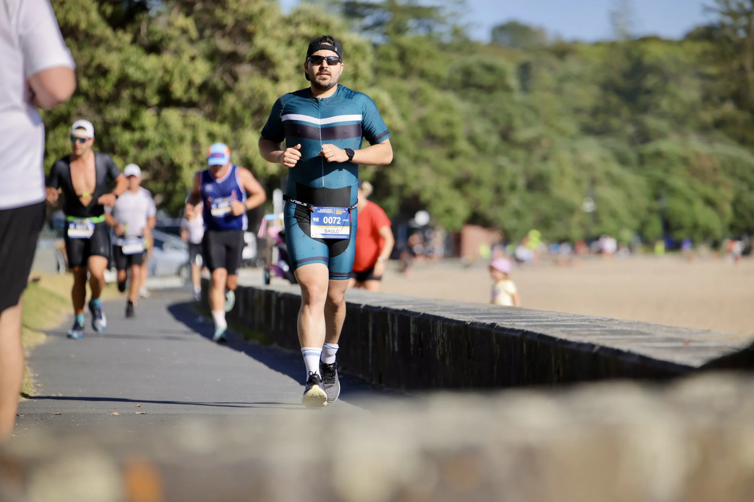A man wearing sunglasses, a blue athletic outfit, and a black cap runs along a paved path during a marathon, with multiple other runners behind him and lush green trees in the background.