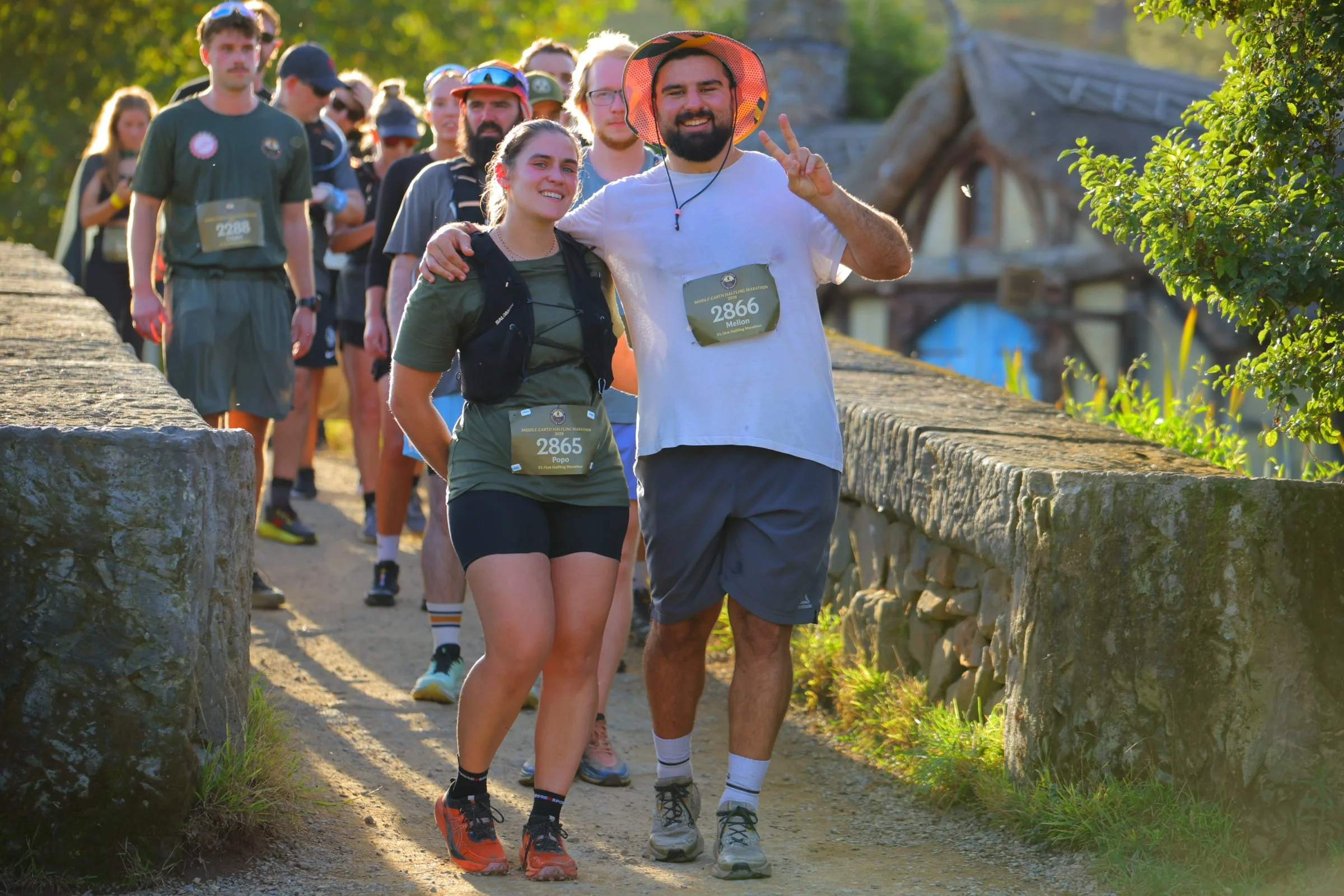 Group of people participating in a marathon, walking along a dirt path, with some smiling and posing for the camera, outdoors in a sunny setting with greenery and a stone bridge in the background.