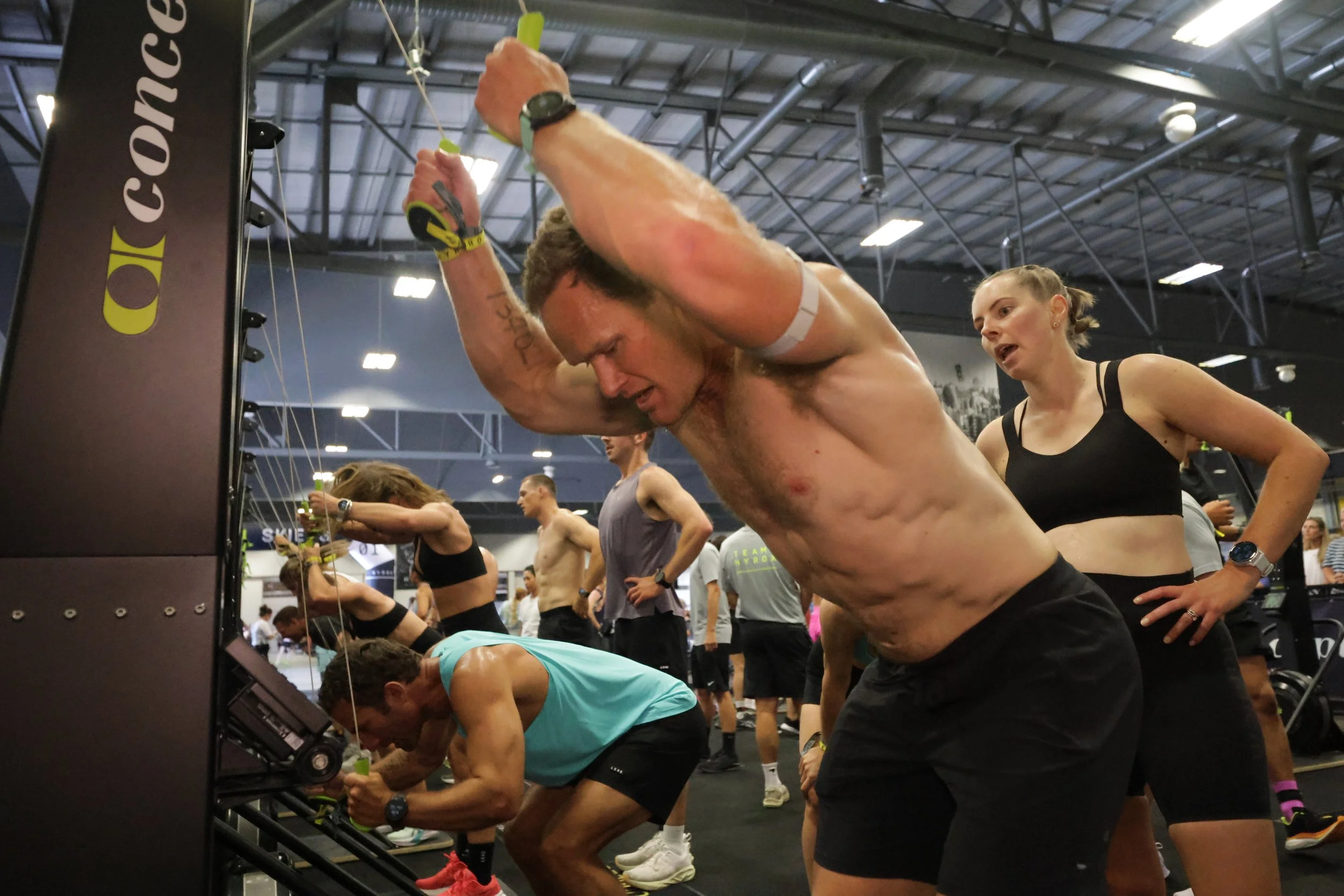 A crowded indoor gym with people participating in a fitness event. A shirtless man is in the foreground, reaching for something, with a woman in a black sports top behind him. Several individuals are seen working out or preparing in the background.