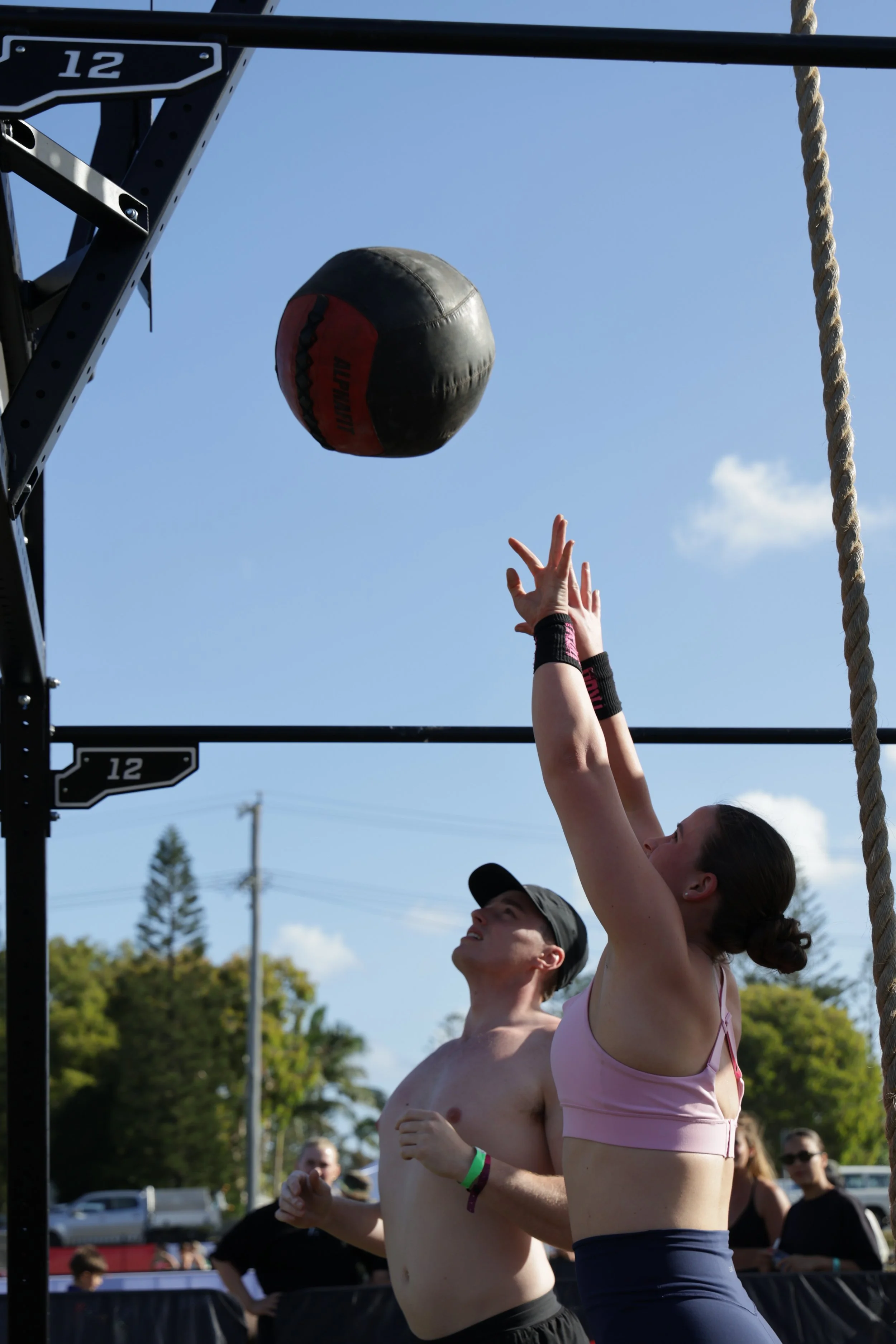 Two people, a woman and a man, participating in an outdoor fitness event. The woman is reaching upward with her hand toward a black and red medicine ball hanging from a rack, and the man stands beside her. The sky is clear with some clouds, and there