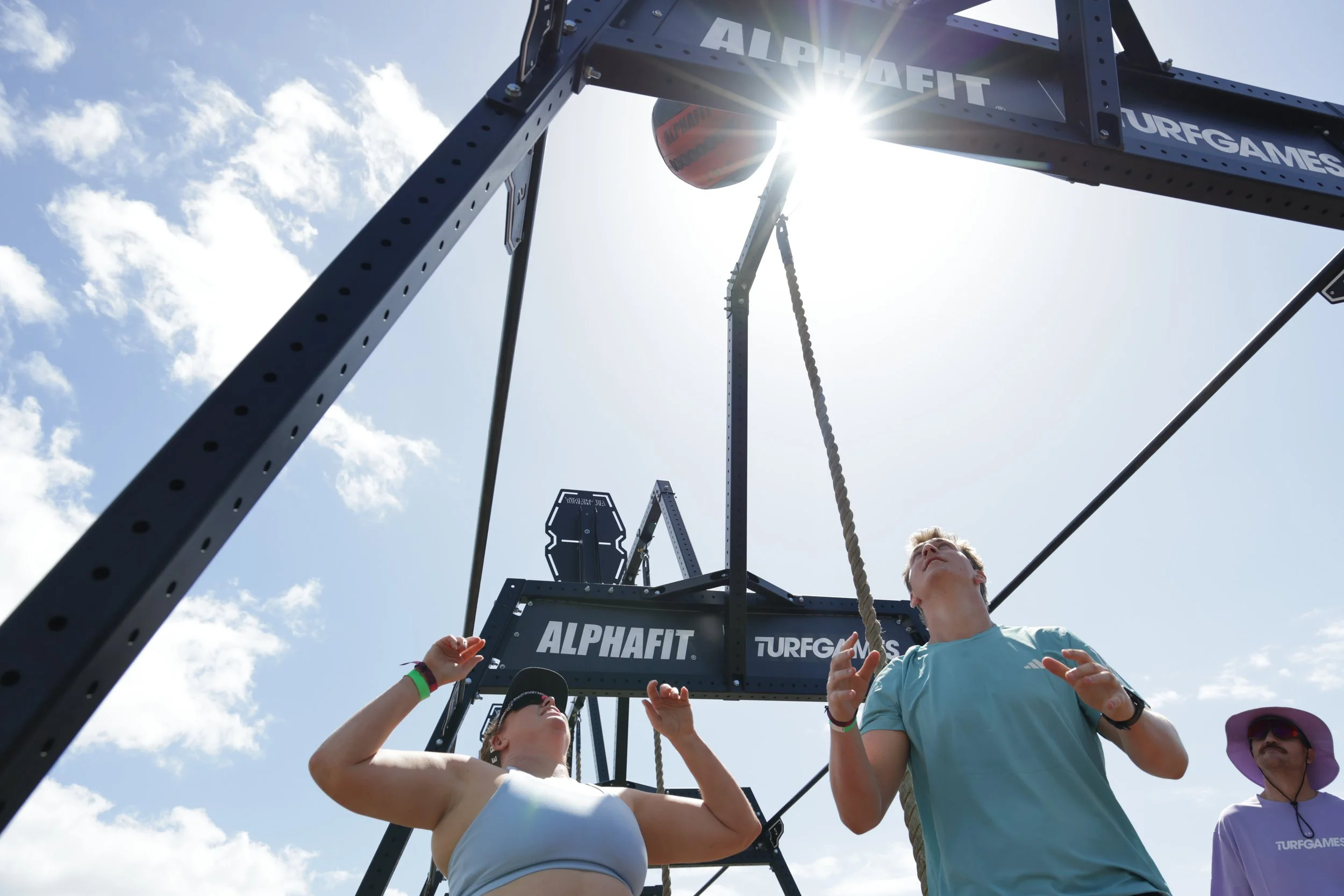 Group of people participating in a tug-of-war competition outdoors under a bright sky with clouds, with a large structure labeled 'ALPHAFIT' and 'TURFGAMES' in the background.