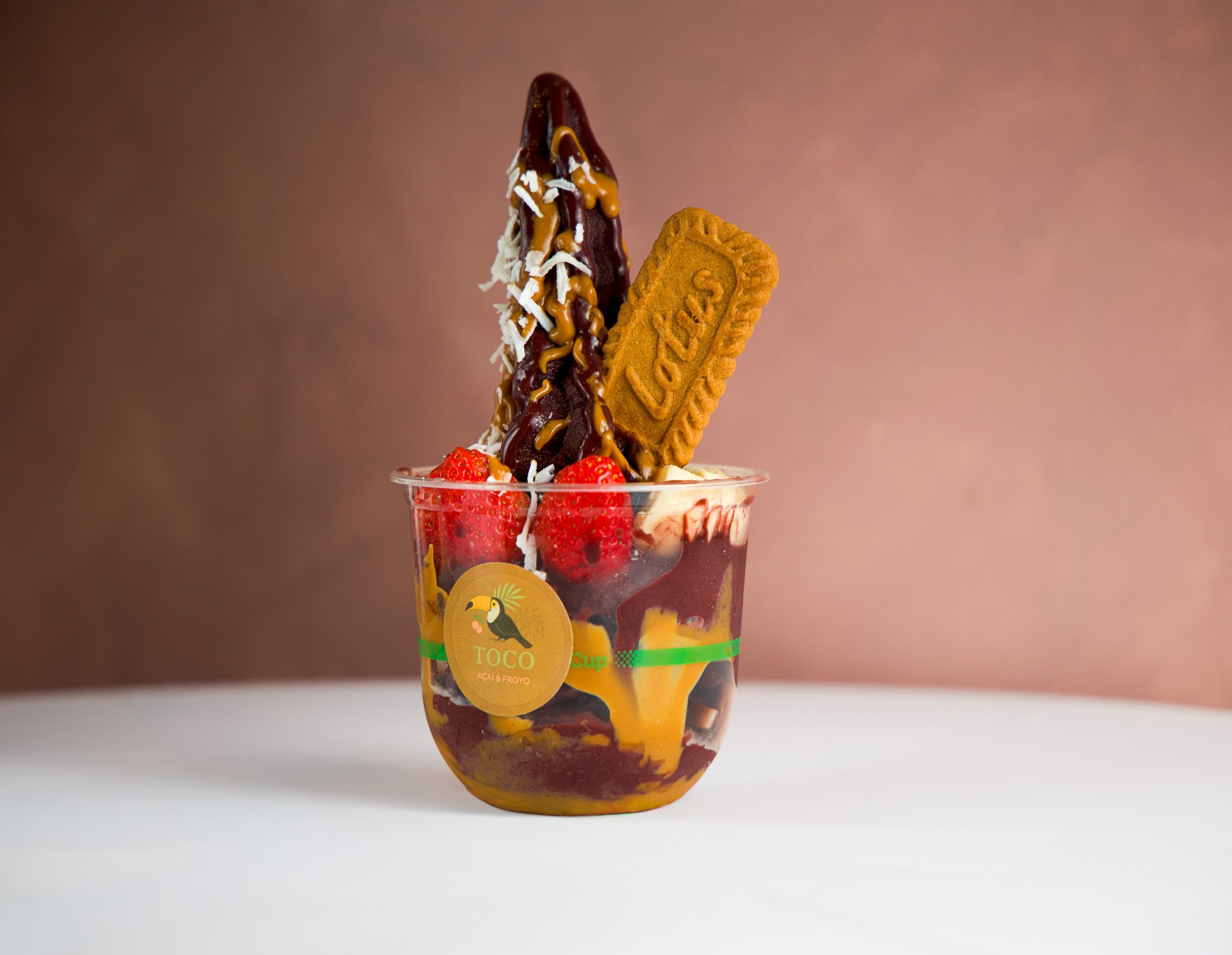 A dessert in a clear cup with strawberries, a cookie, chocolate syrup, and a yellow sauce. The cup has a label with a toucan and the words "TOCO Açaí & Froyo."