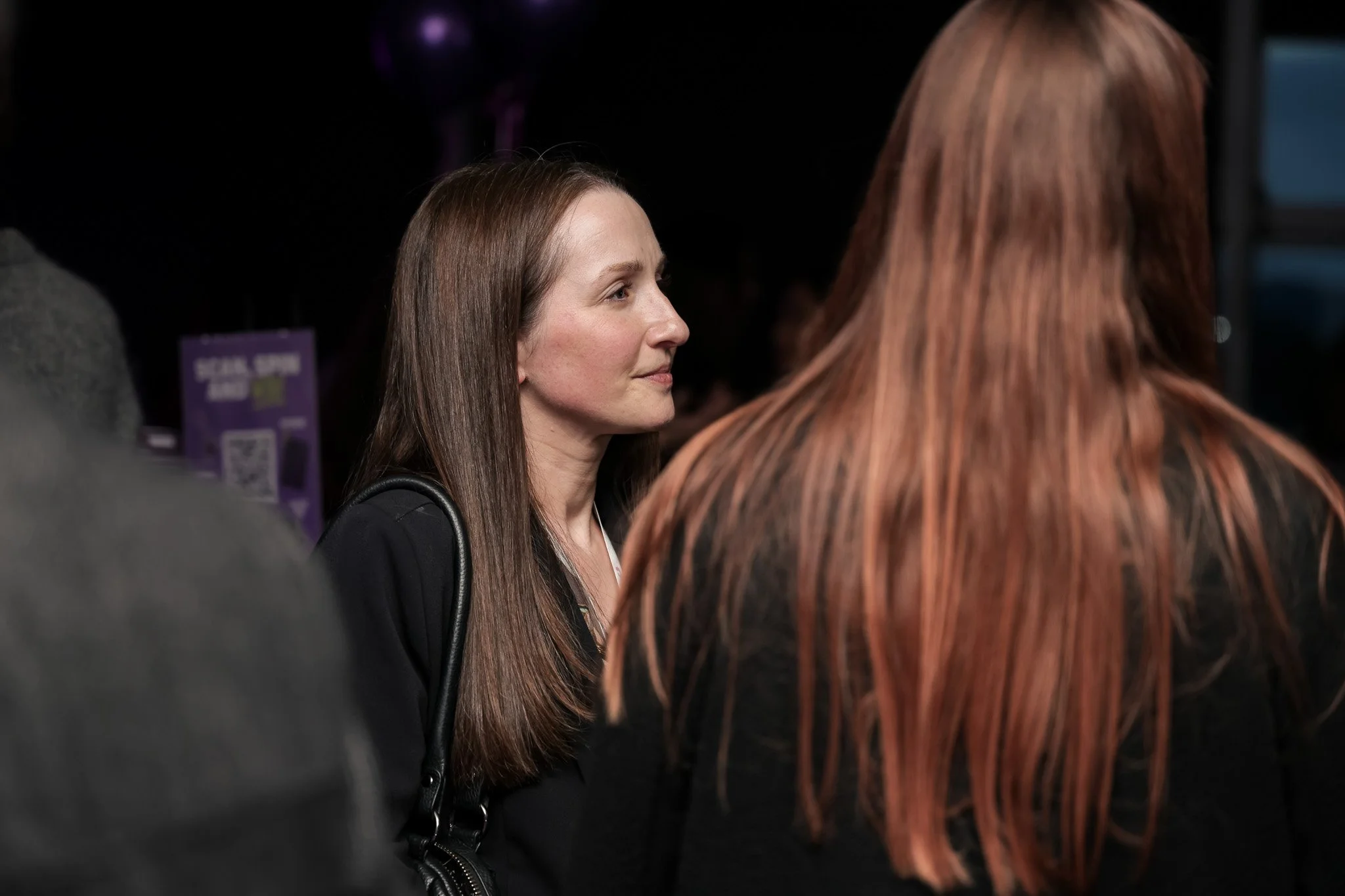 Two women with brown and red hair engaged in conversation at a conference or event, with a purple informational sign in the background.
