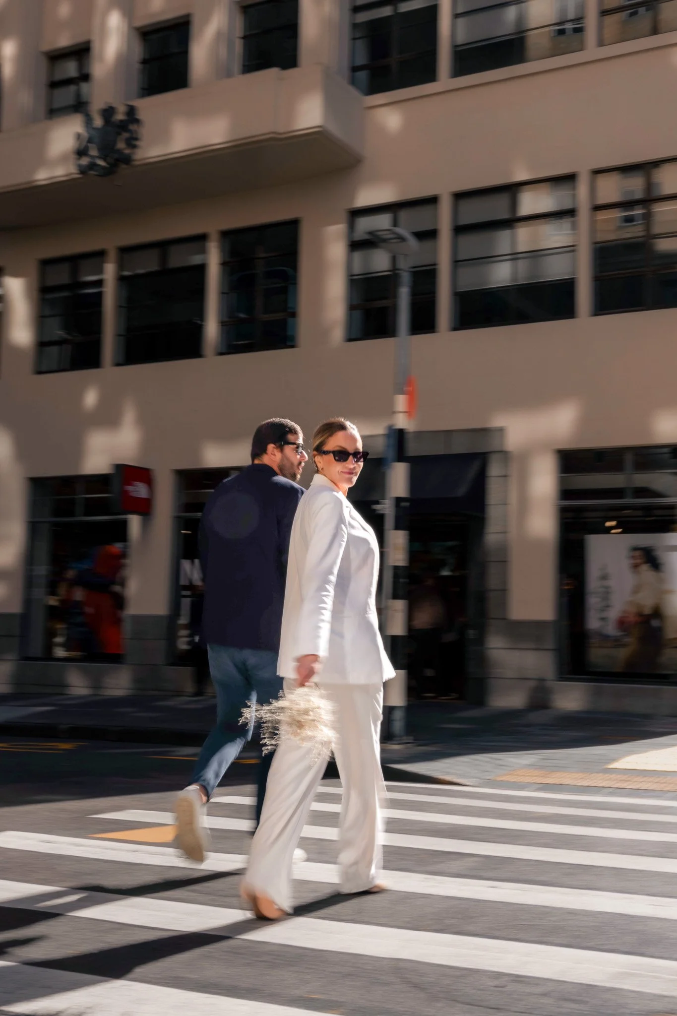 A woman in a white suit and sunglasses walking across a city street crosswalk with a man in a dark jacket and jeans. Modern building facade in the background.