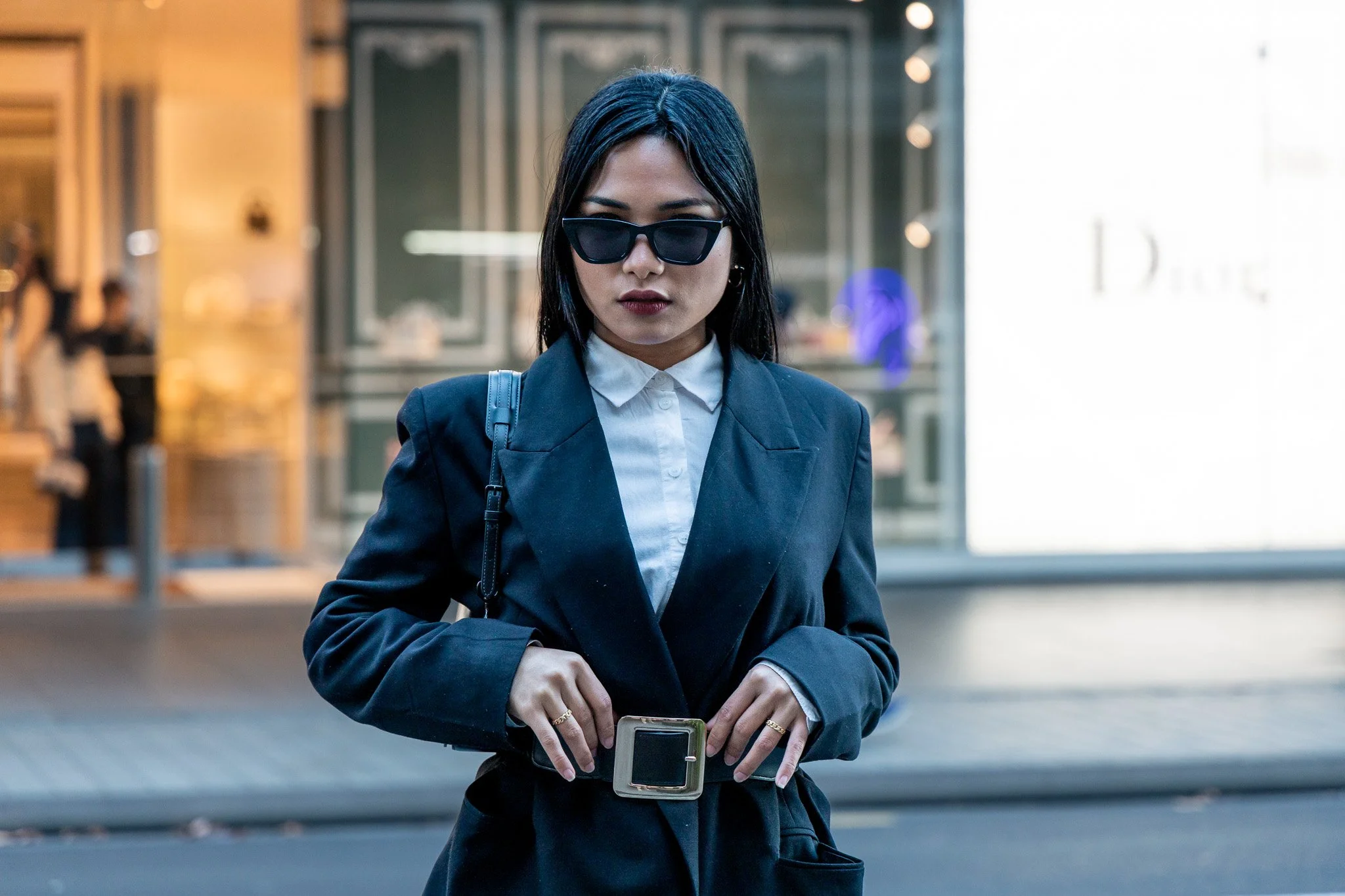 A woman with dark hair wearing sunglasses, a white shirt, and a dark blazer, looking down at her phone in an urban setting.