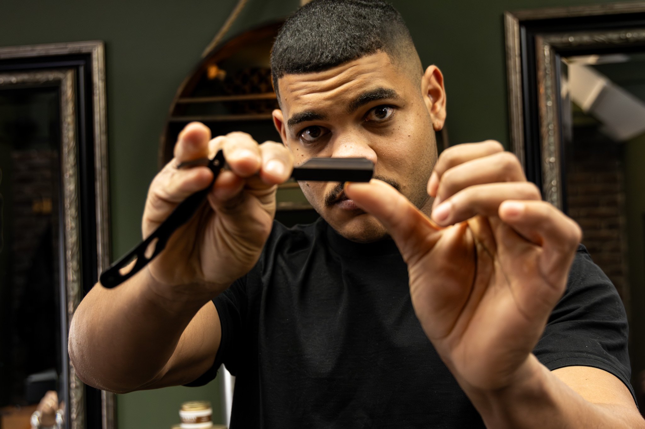 A man with short, dark hair and a black shirt is focused on shaving his beard with an electric razor in a room with green walls and framed mirrors.