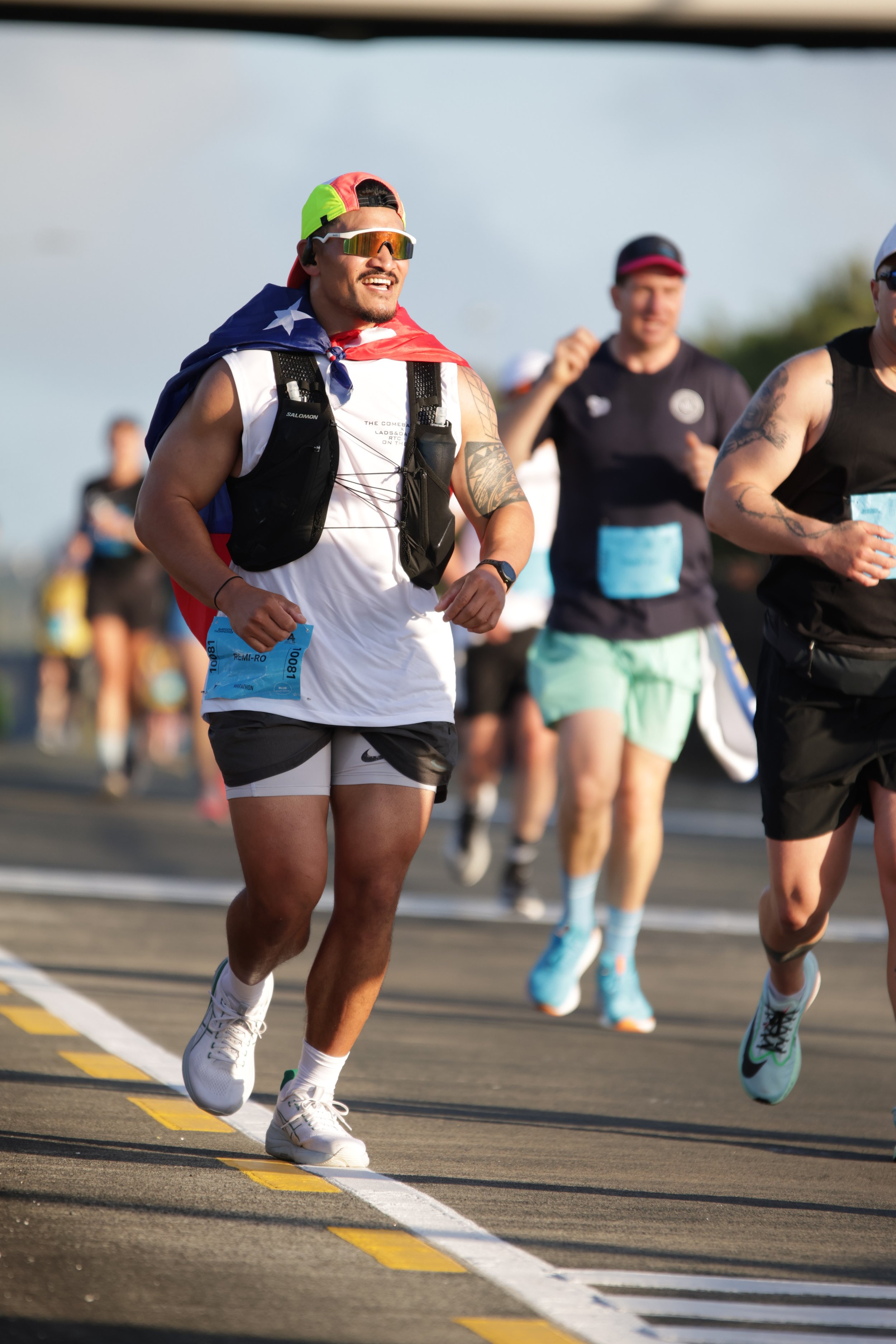 People running in a race, with a man in a white tank top, black shorts, white shoes, wearing sunglasses, a backpack, and a colorful cap, leading the group.