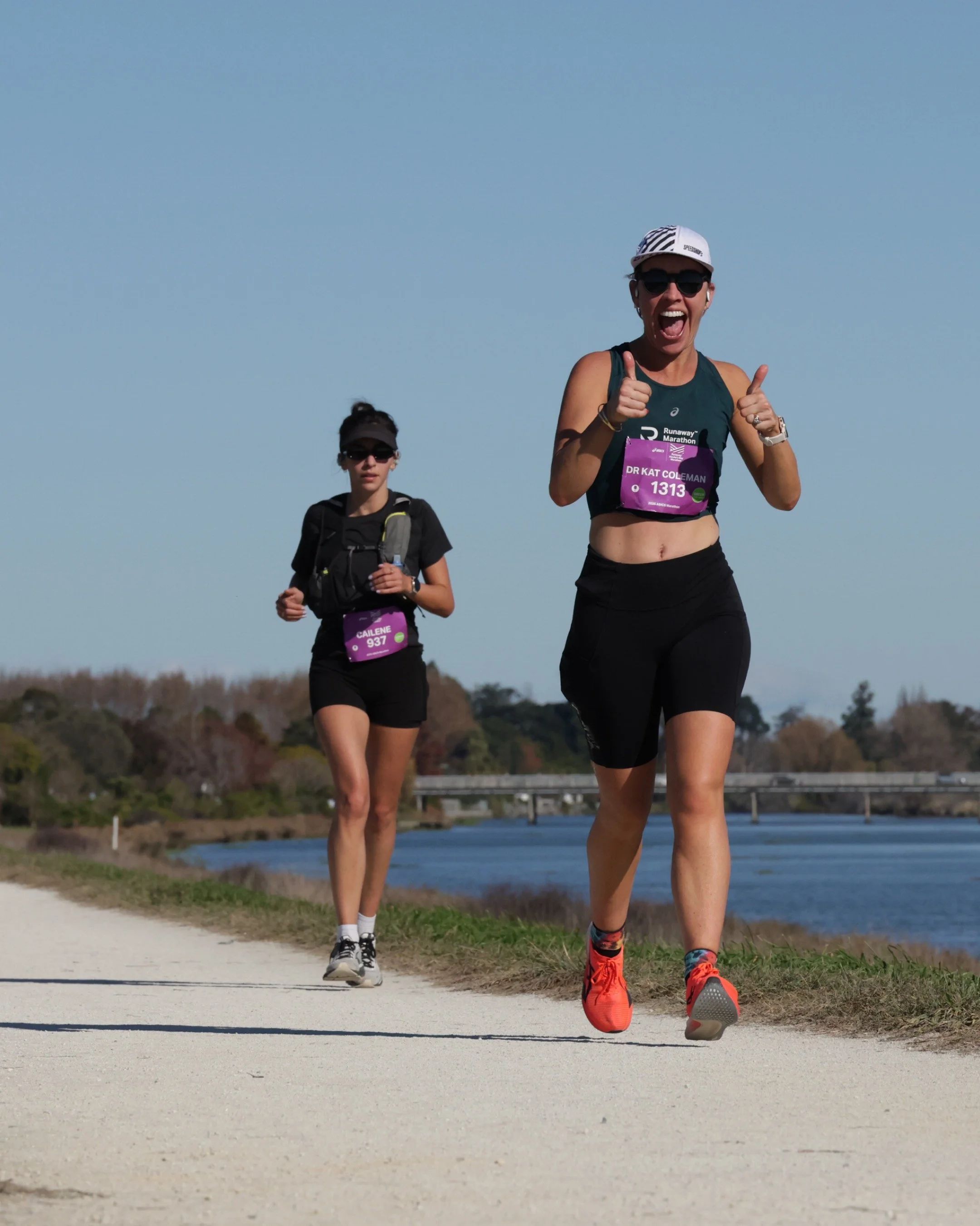 Two women running along a path beside a body of water, with a bridge in the background, under a clear blue sky.