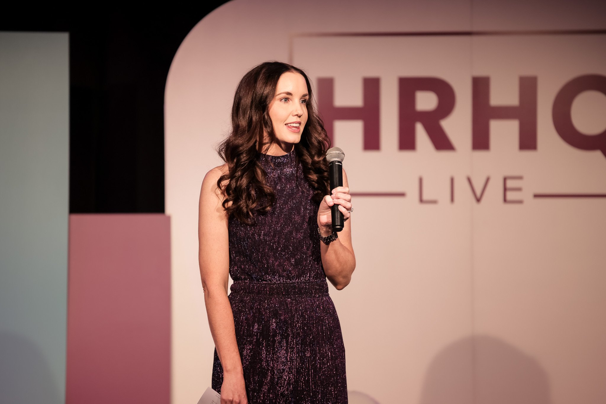 A woman with long, dark, wavy hair speaking into a microphone on stage. She is wearing a sleeveless, glittery, dark purple dress. Behind her, part of a sign with the letters "HRH" is visible.