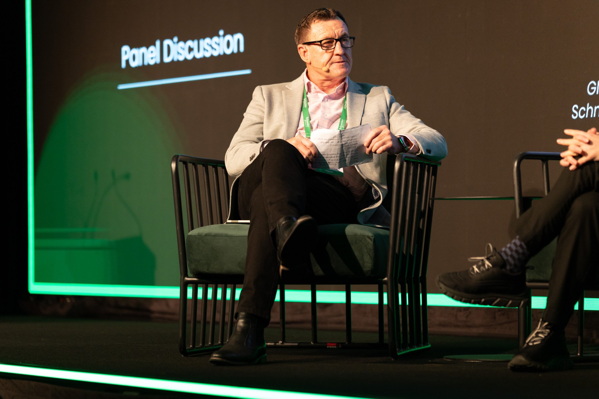 A man wearing glasses, a gray blazer, and a pink shirt sitting on a chair during a panel discussion at a conference.