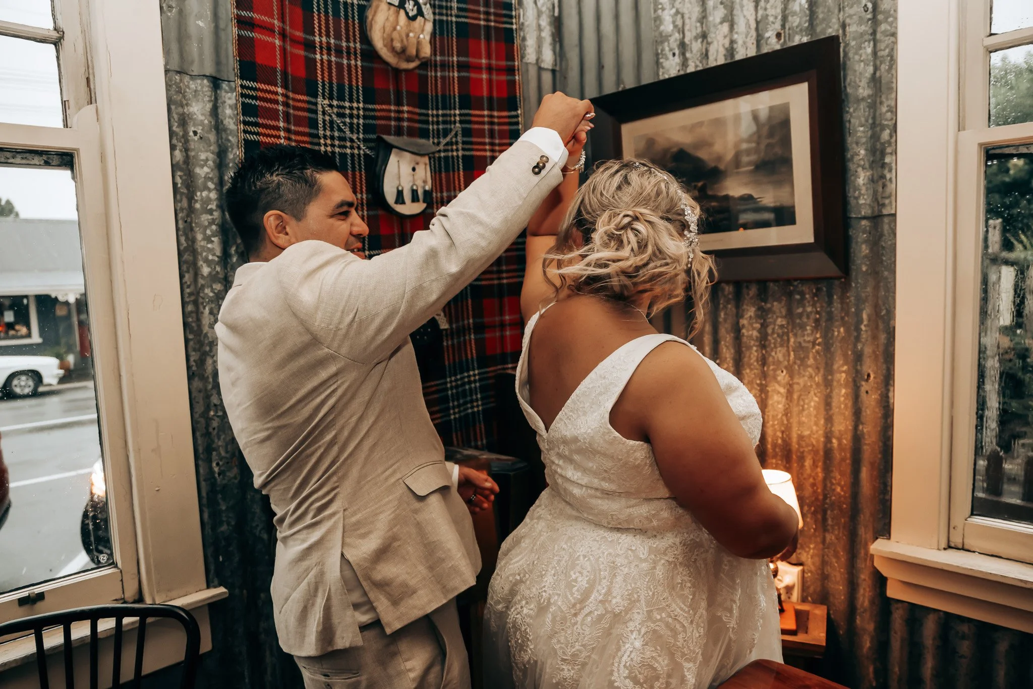 A groom and bride are dancing together inside a room with rustic walls and curtains, near a window. The groom is in a light gray suit, and the bride is in a white lace wedding dress with curly hair and floral hair accessories.