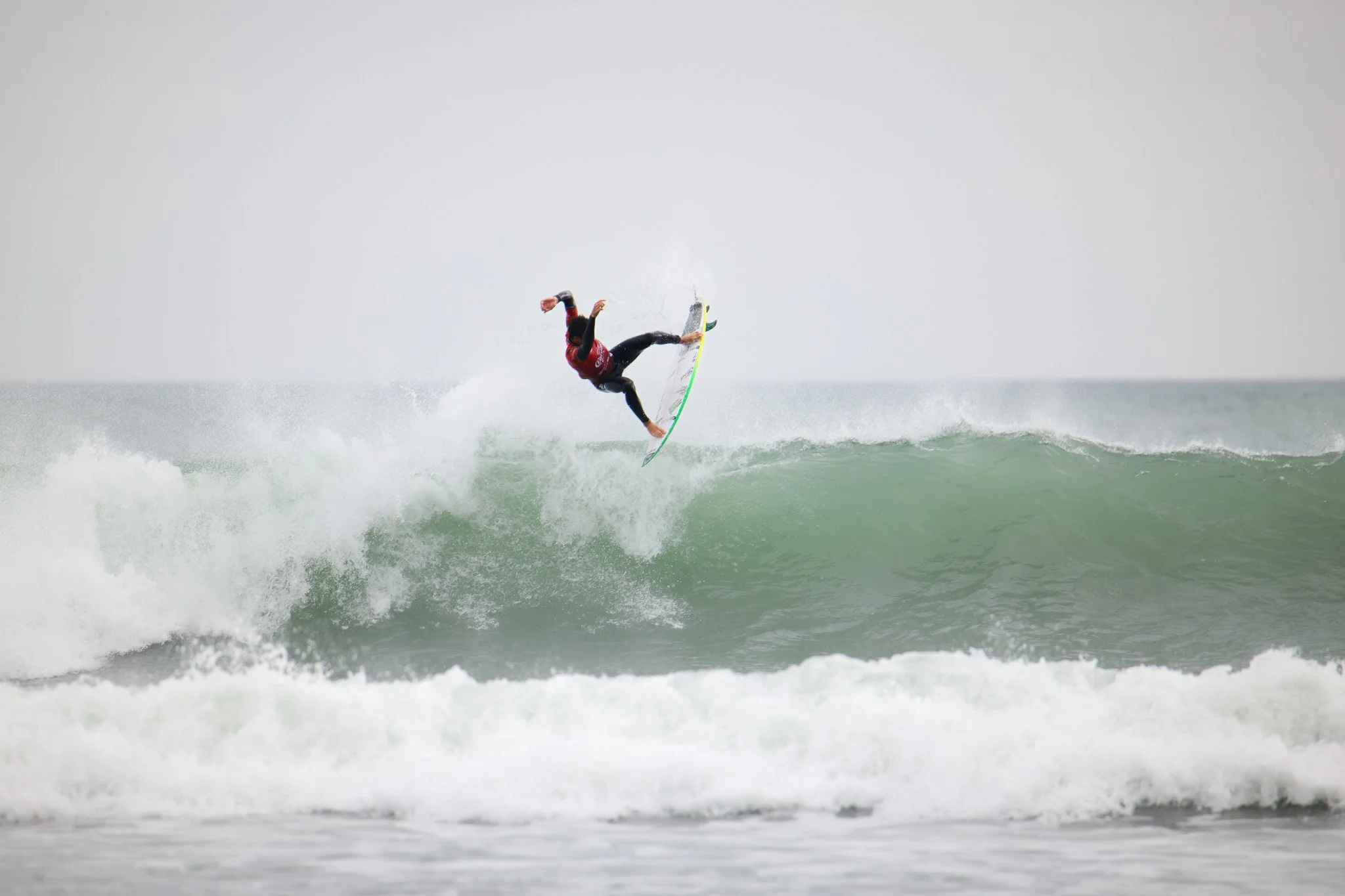 A person surfing on a wave in the ocean, wearing a red shirt and black wetsuit, performing an aerial maneuver.