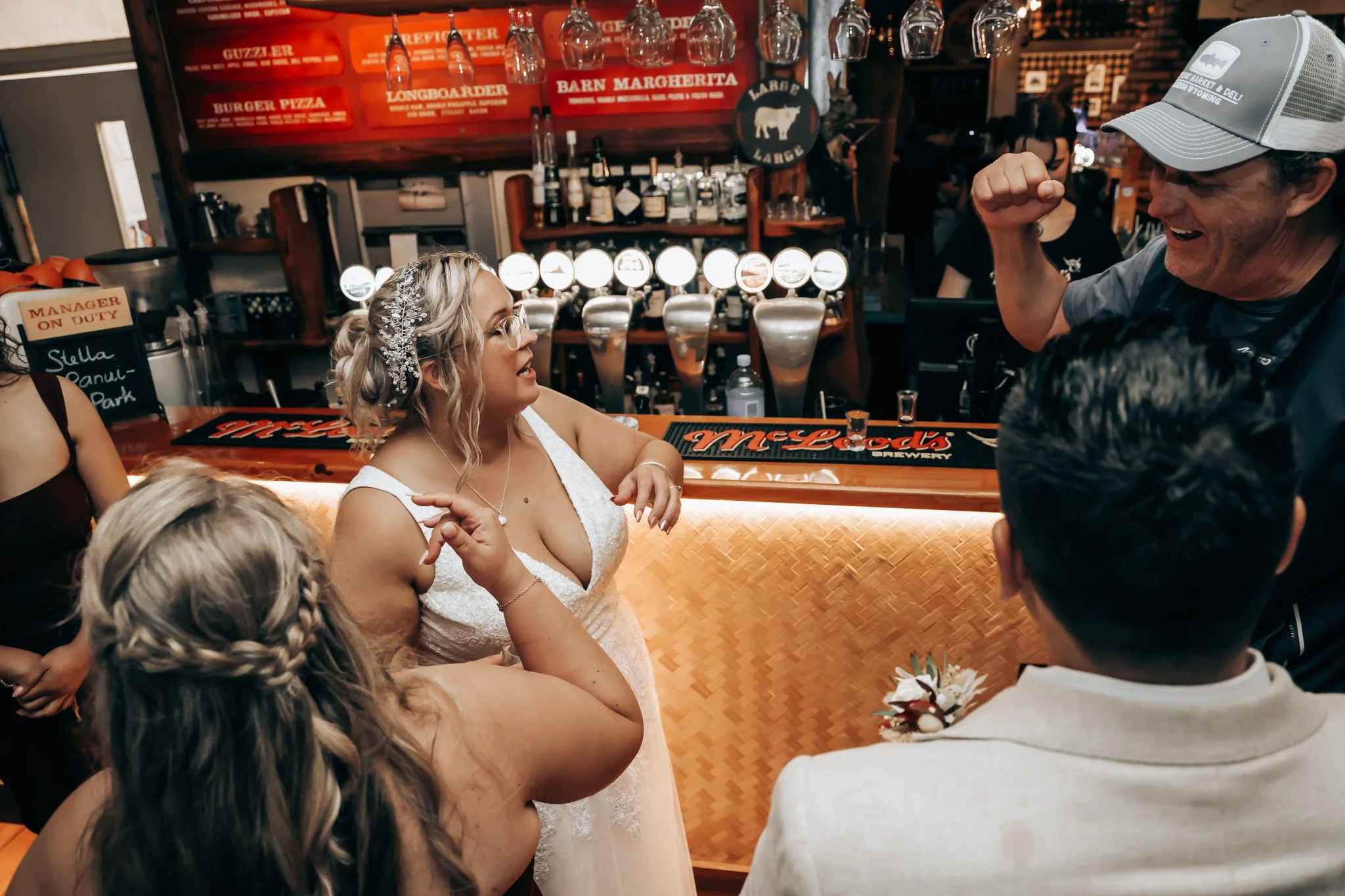 A wedding reception scene at a bar with a bride in a white lace wedding dress and a man, possibly the groom, in a white shirt. They appear to be talking and enjoying themselves. The bar has various bottles and taps, with people in the background.