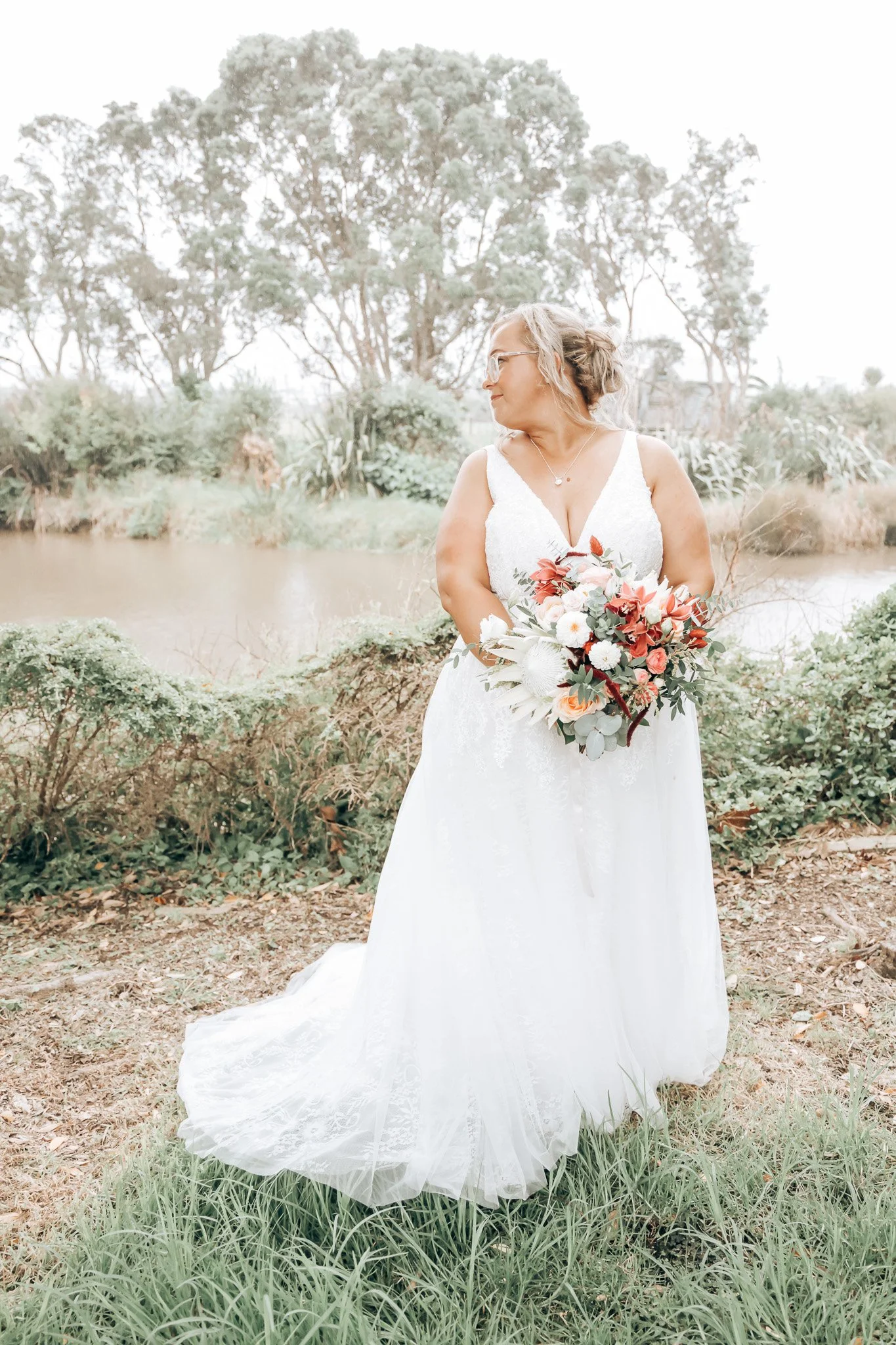 A woman in a white wedding dress holding a bouquet of flowers, standing outdoors near a body of water with trees in the background.
