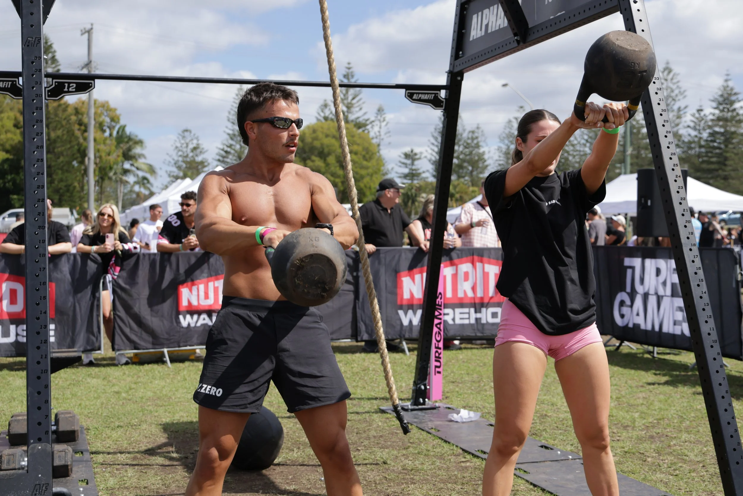 A shirtless man wearing sunglasses holding a kettlebell and a woman in a black shirt and pink shorts lifting a kettlebell at a fitness competition outdoors with spectators watching.