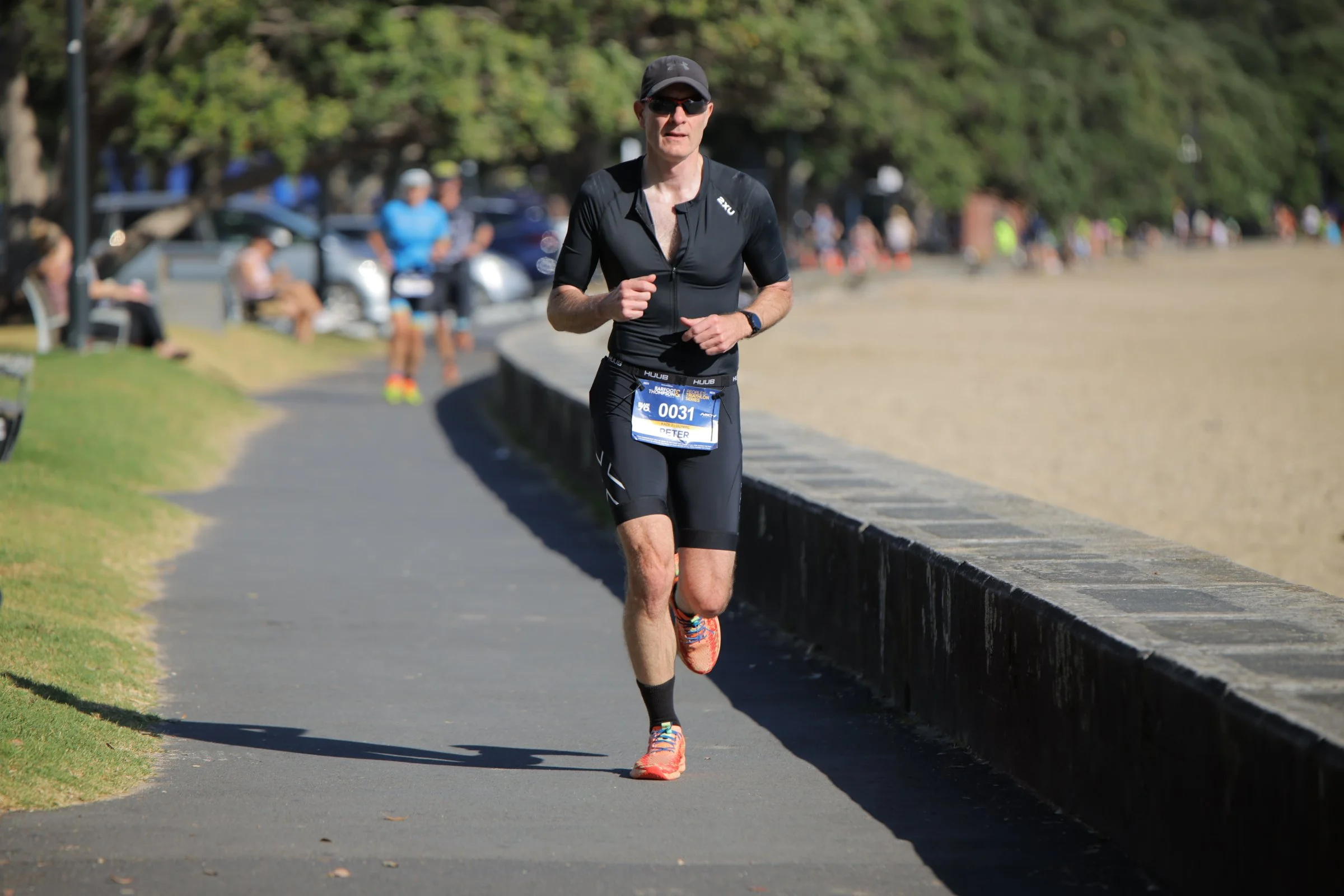 A man running on a paved path near a grassy area during a race or marathon, with trees, cars, and onlookers in the background.