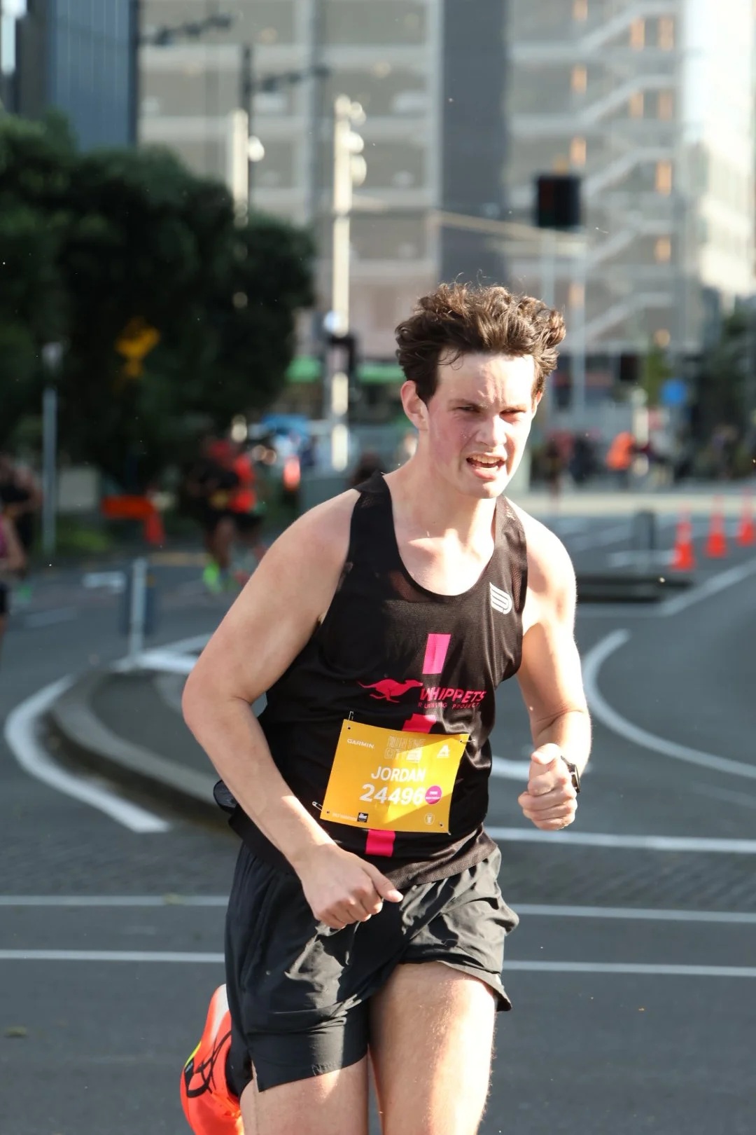 A male marathon runner with short curly hair running outdoors on a city street, wearing a black sleeveless shirt with a pink logo, black shorts, and a yellow race bib with the name Jordan and number 24496.