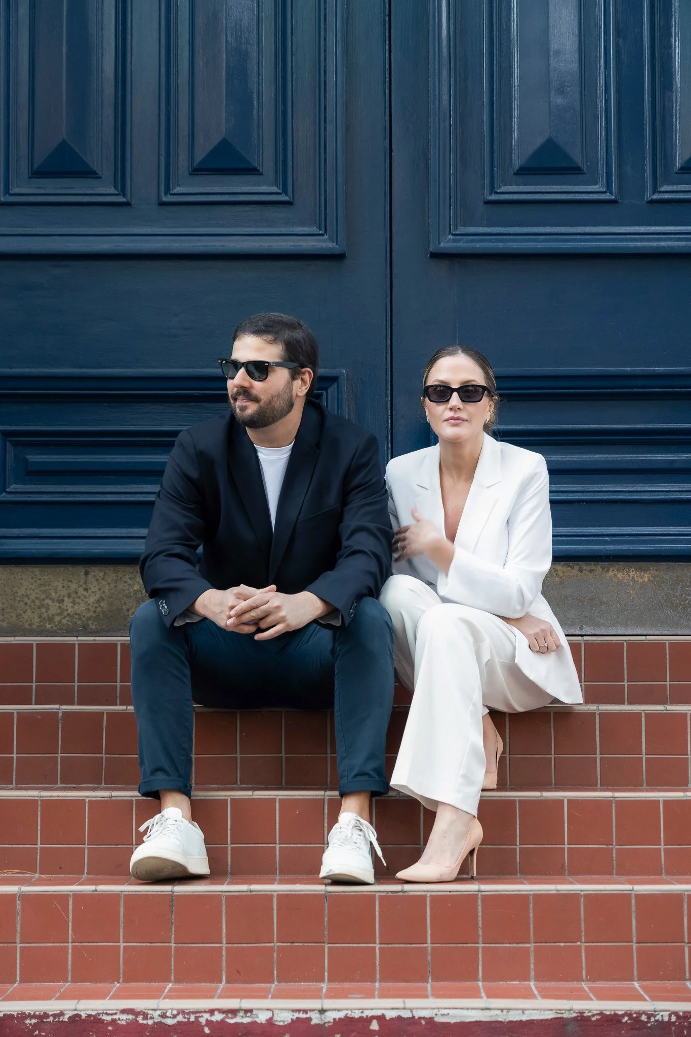 A man and a woman sitting on steps against a large blue door, both wearing sunglasses. The man is in a dark suit with white sneakers, while the woman is in a white suit with nude heels.