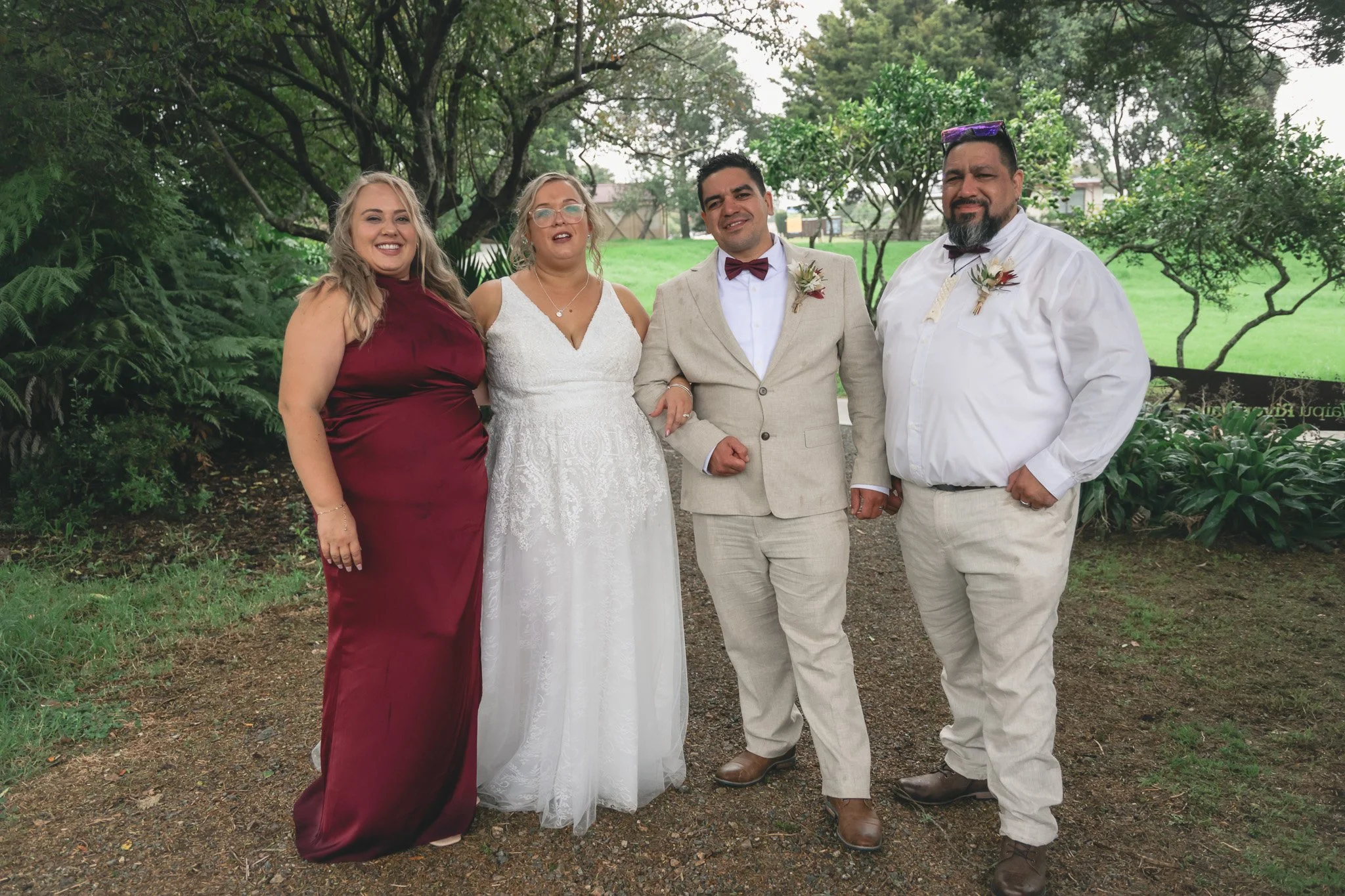 Group of five people standing outdoors in a garden or park, dressed in formal and semi-formal attire, with trees and greenery in the background.