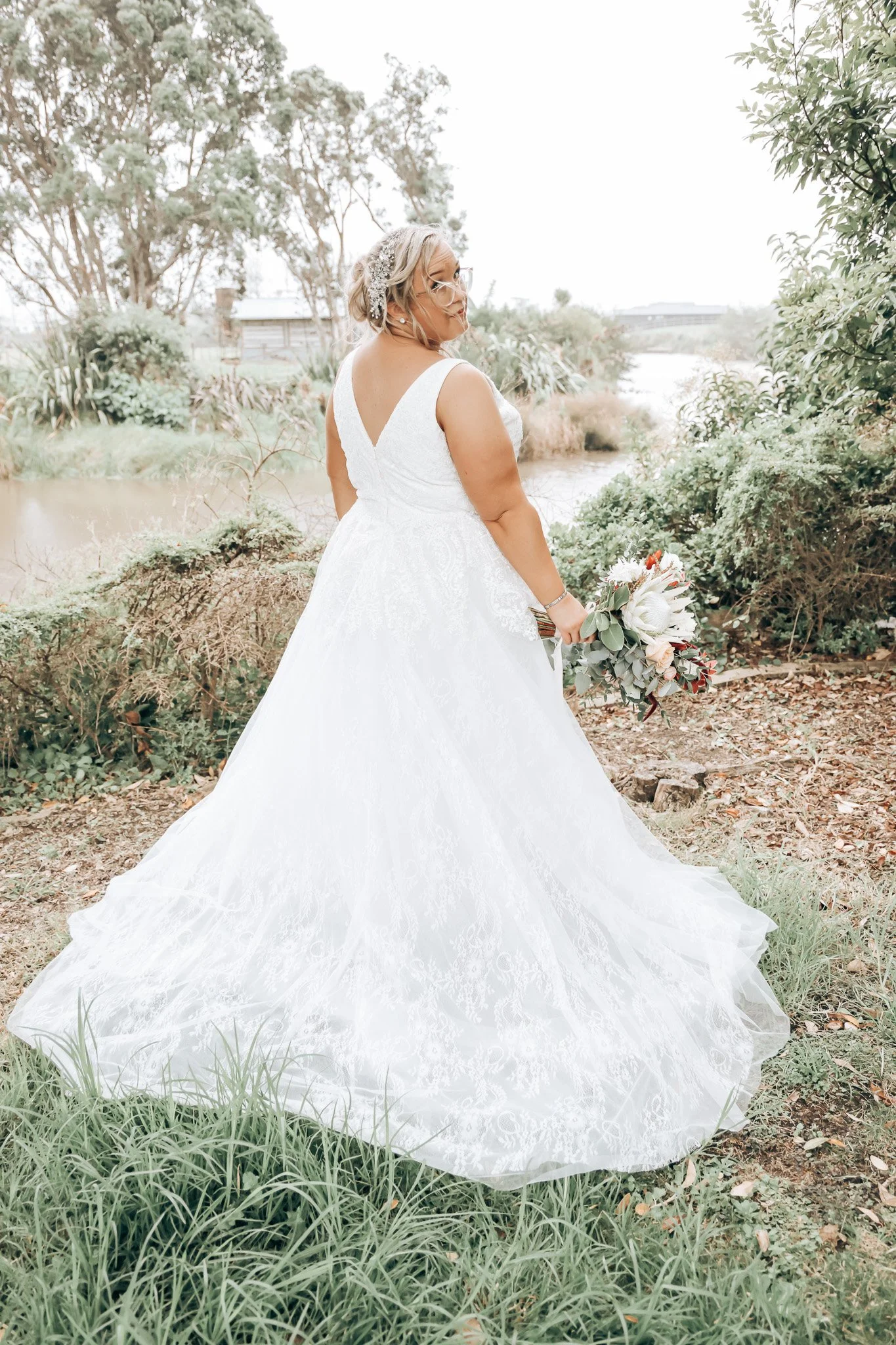 A bride in a white wedding dress holding a bouquet of flowers outdoors in a natural setting near a body of water, with trees and shrubs in the background.