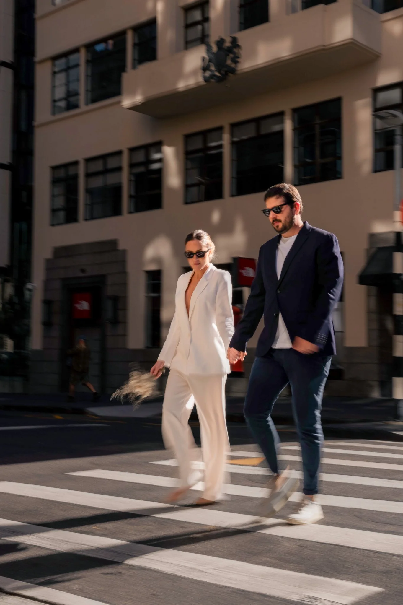 A man and woman holding hands and crossing a city street on a crosswalk, both wearing sunglasses, with the woman dressed in a white suit and the man in a dark suit, as sunlight casts shadows on a modern building in the background.