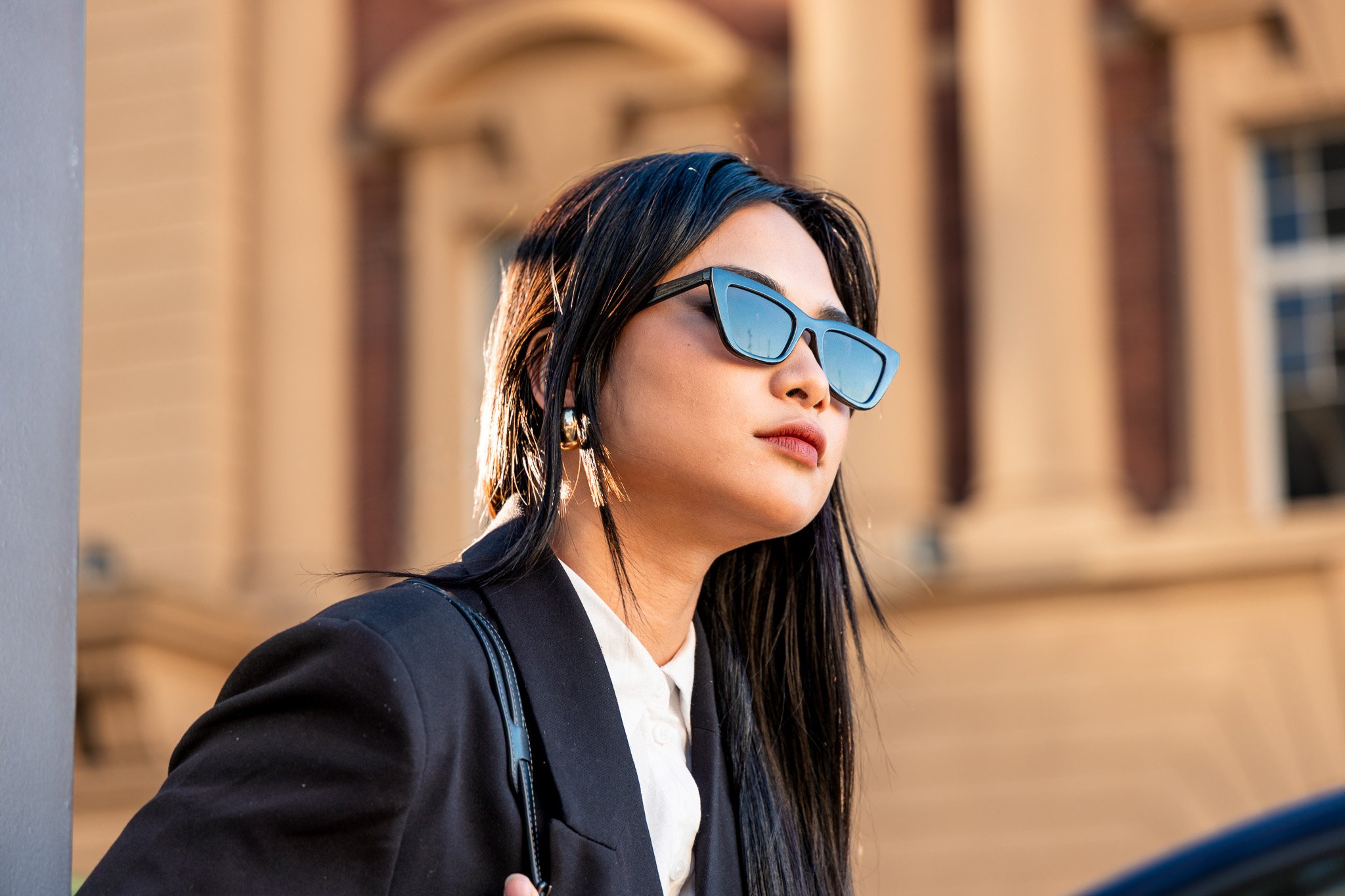 A woman with long black hair wearing sunglasses, a black blazer, a white shirt, and gold earrings. She is standing outdoors with a building in the background.