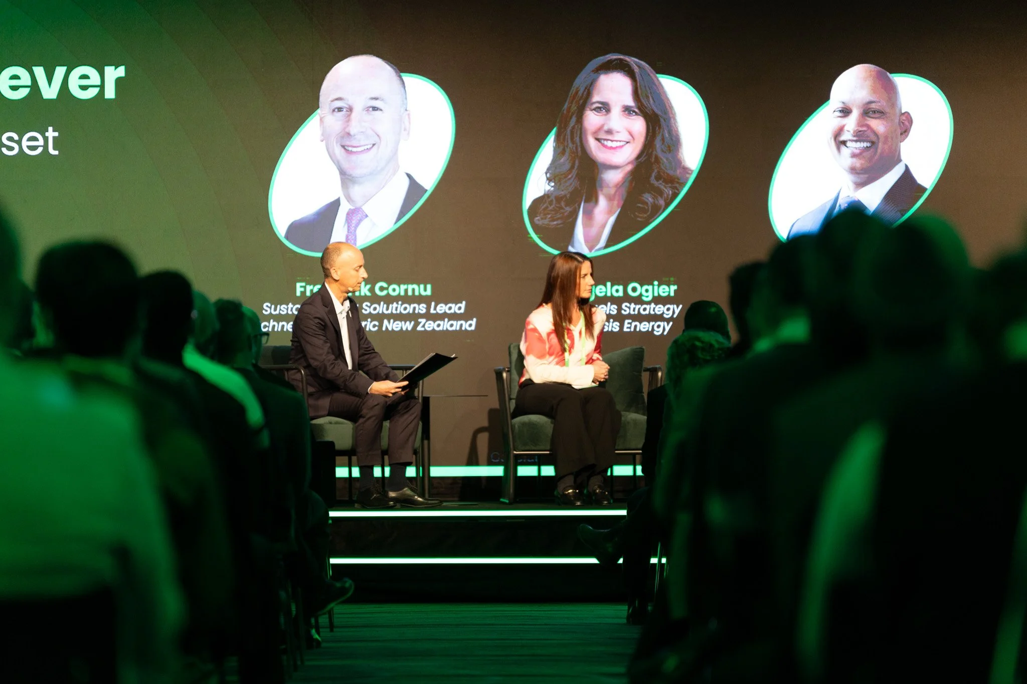 Photo of a conference or event with people sitting and two individuals on stage in front of a large screen showing headshots of three people and their names and titles.