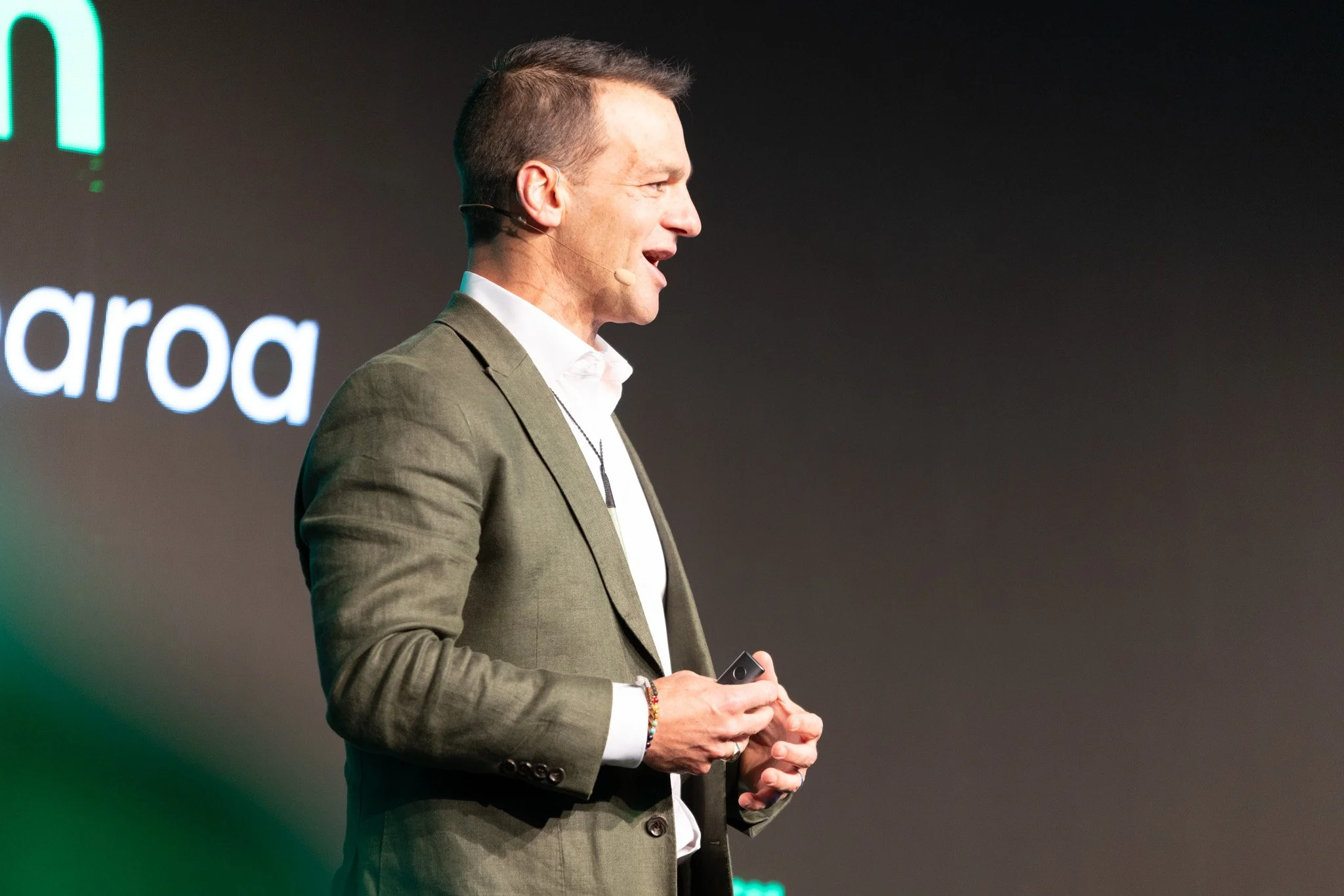 A man in a gray suit giving a presentation on stage, with a microphone headset, holding a remote in his hand, standing in front of a dark background with partially visible illuminated text.