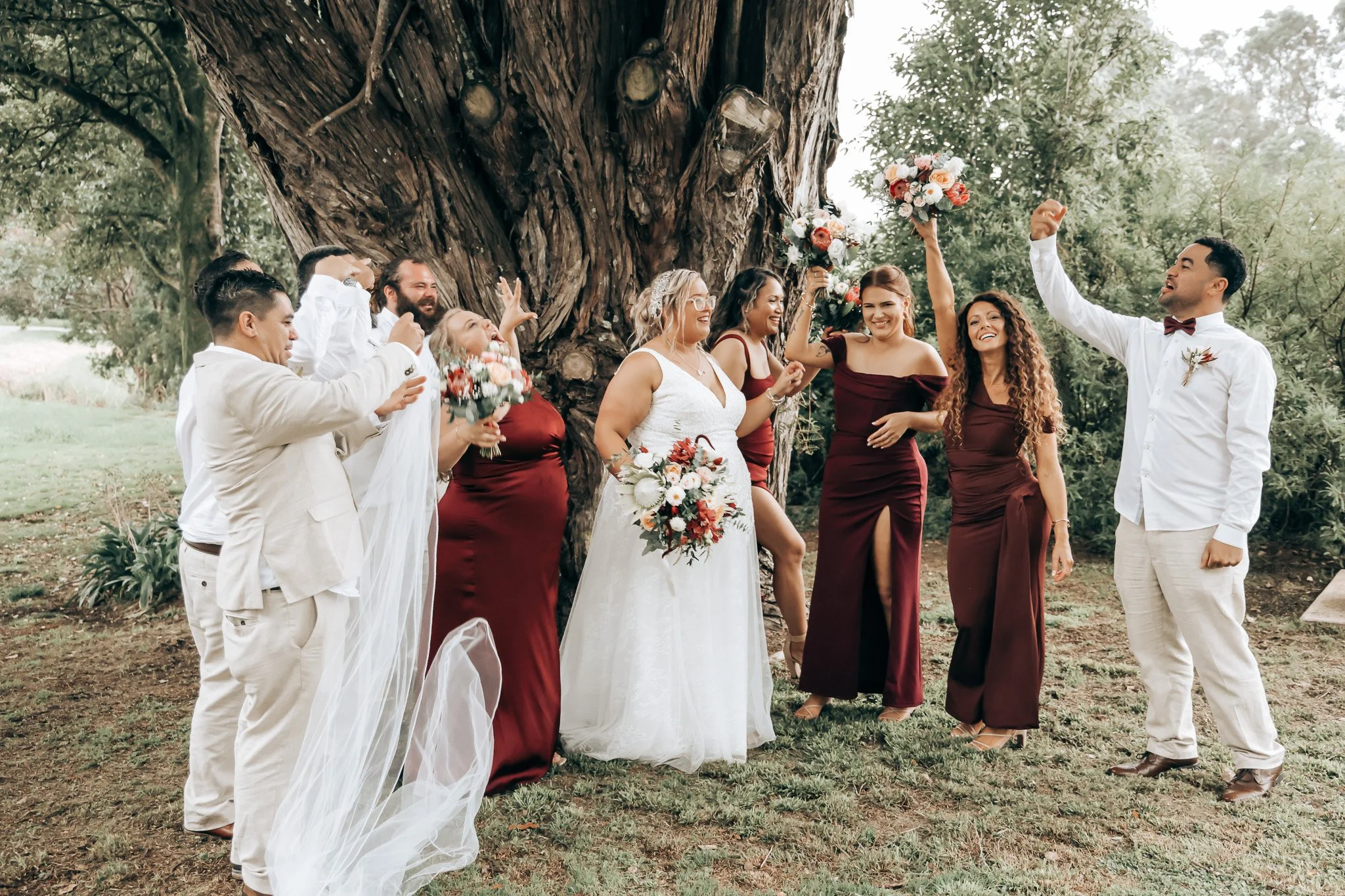 A wedding celebration outdoors with a bride, groom, and friends standing under a large tree, holding bouquets of flowers, and smiling.