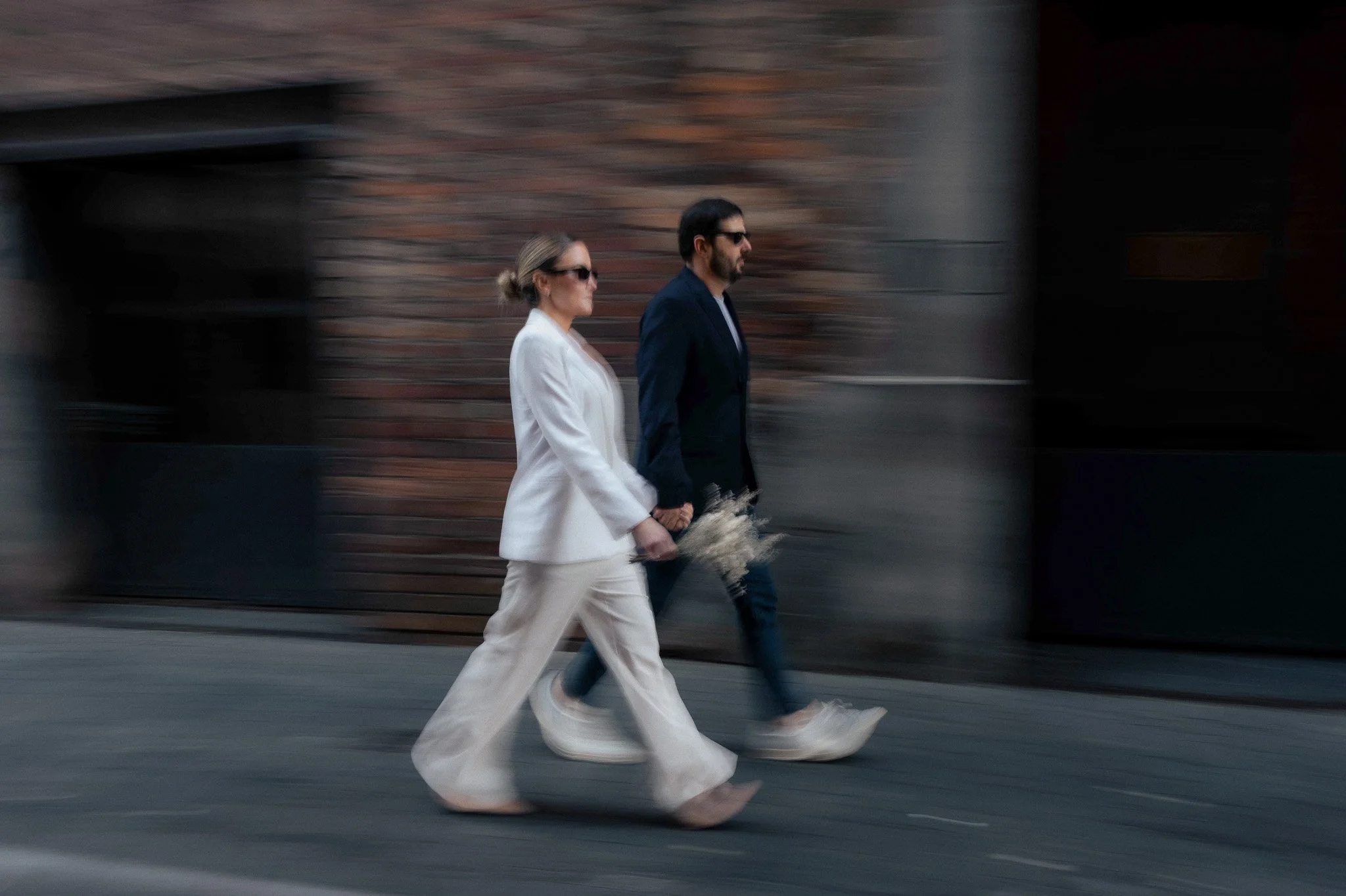 A man and woman walking together on the sidewalk, holding hands, with motion blur background showing a brick wall.