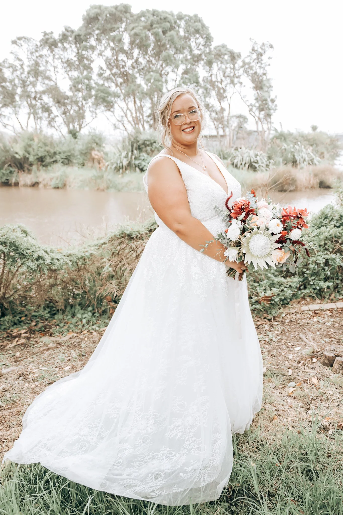 A woman in a white wedding dress holding a bouquet of flowers, standing outdoors near a body of water with trees in the background.