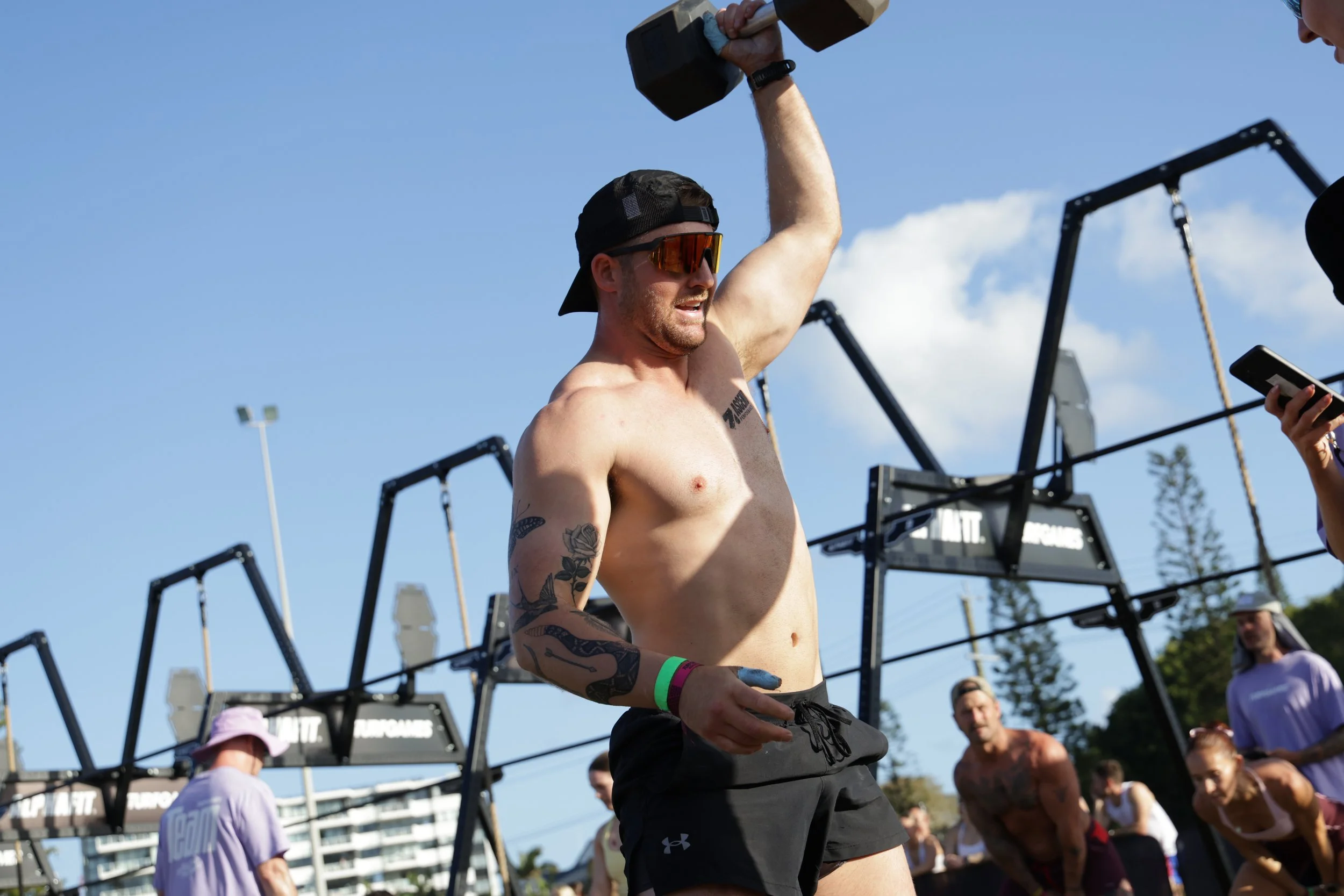 A shirtless man wearing sunglasses, a black cap backward, and black shorts lifting a dumbbell outdoors with a crowd watching. The scene includes gym equipment and a bright blue sky.