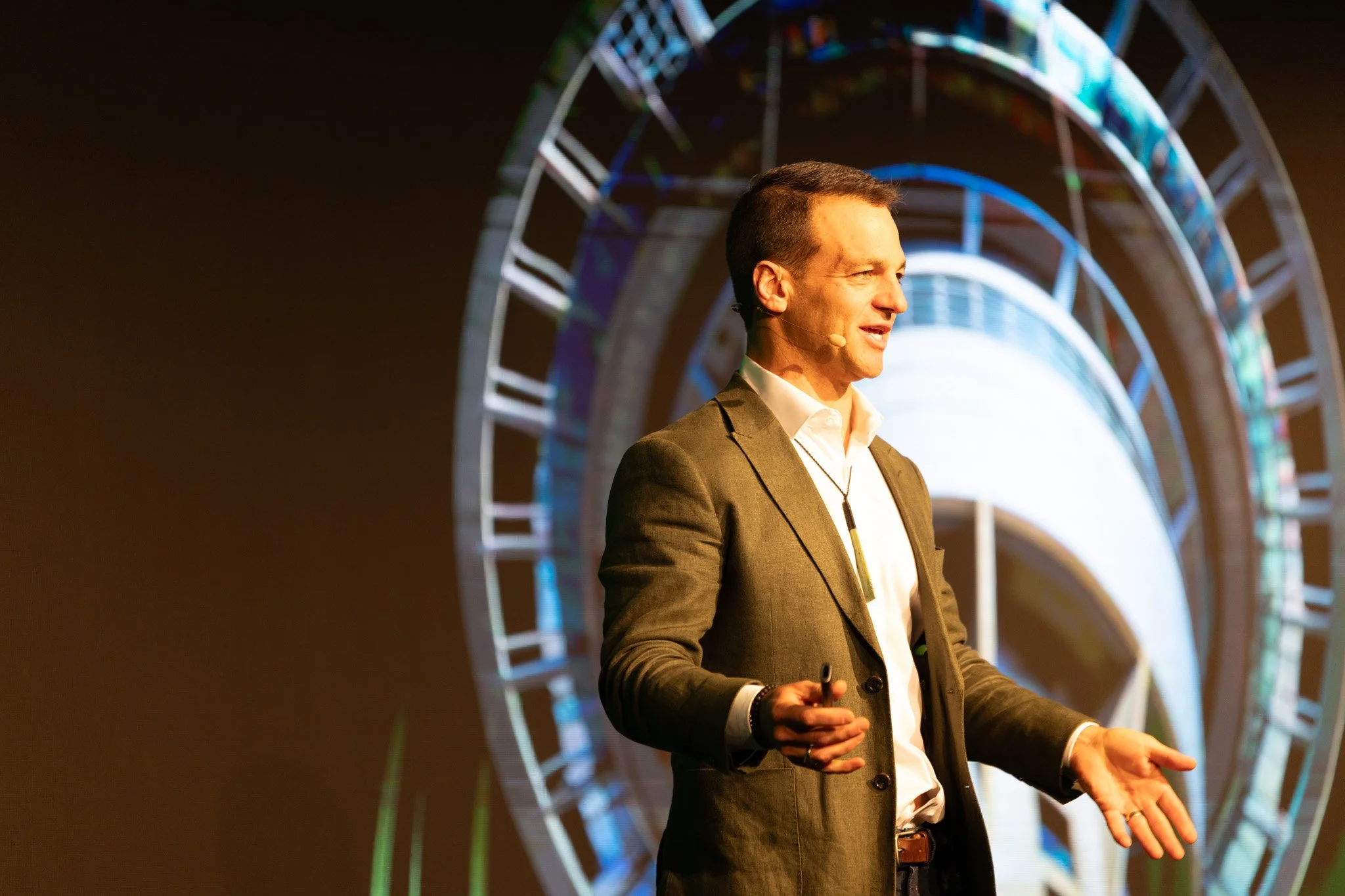 A man in a suit speaking at an event with a large, illuminated Ferris wheel in the background.