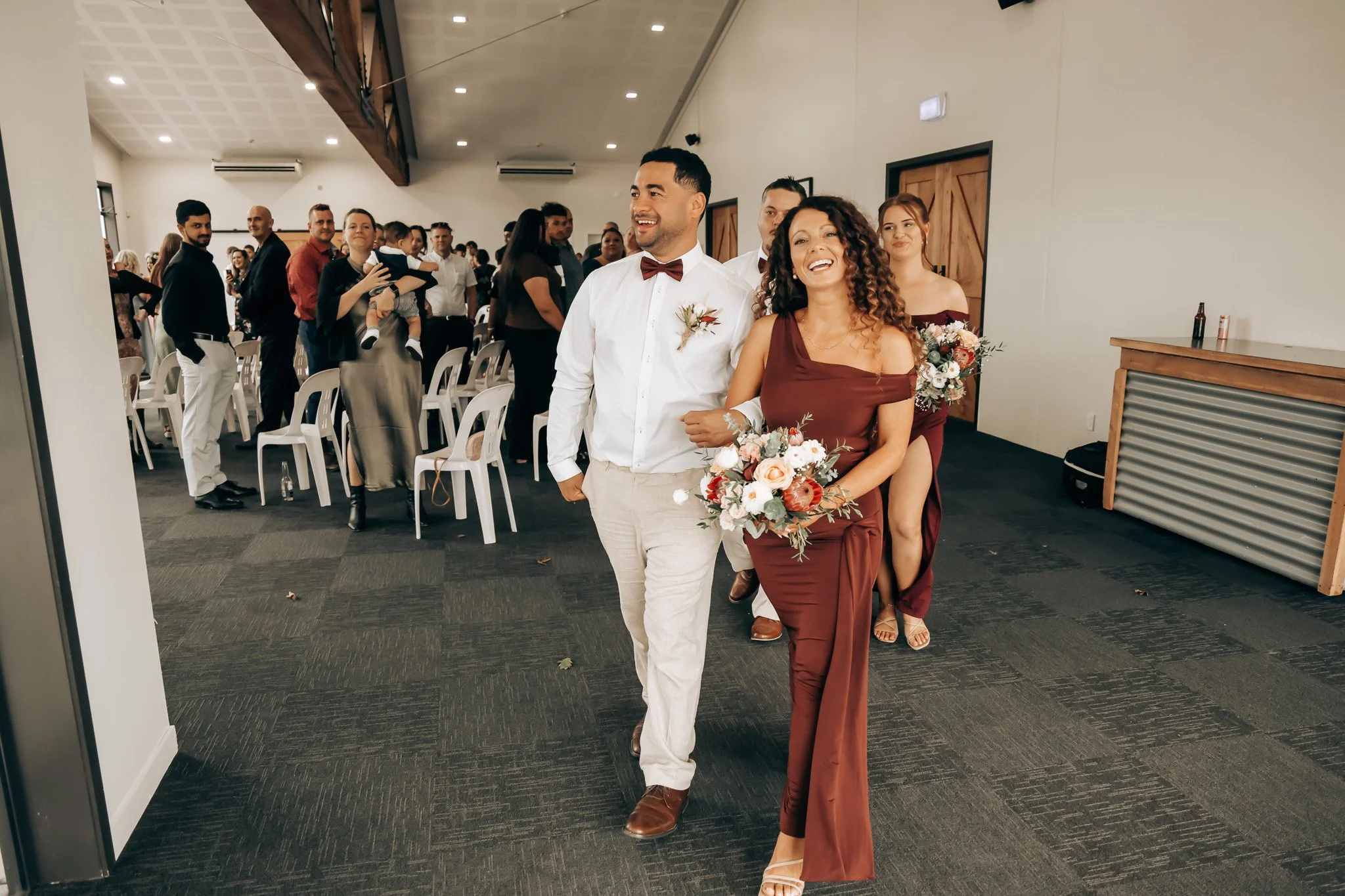 Couple walking down the aisle at a wedding reception, smiling and holding flowers, surrounded by guests seated and standing in a large, well-lit room.