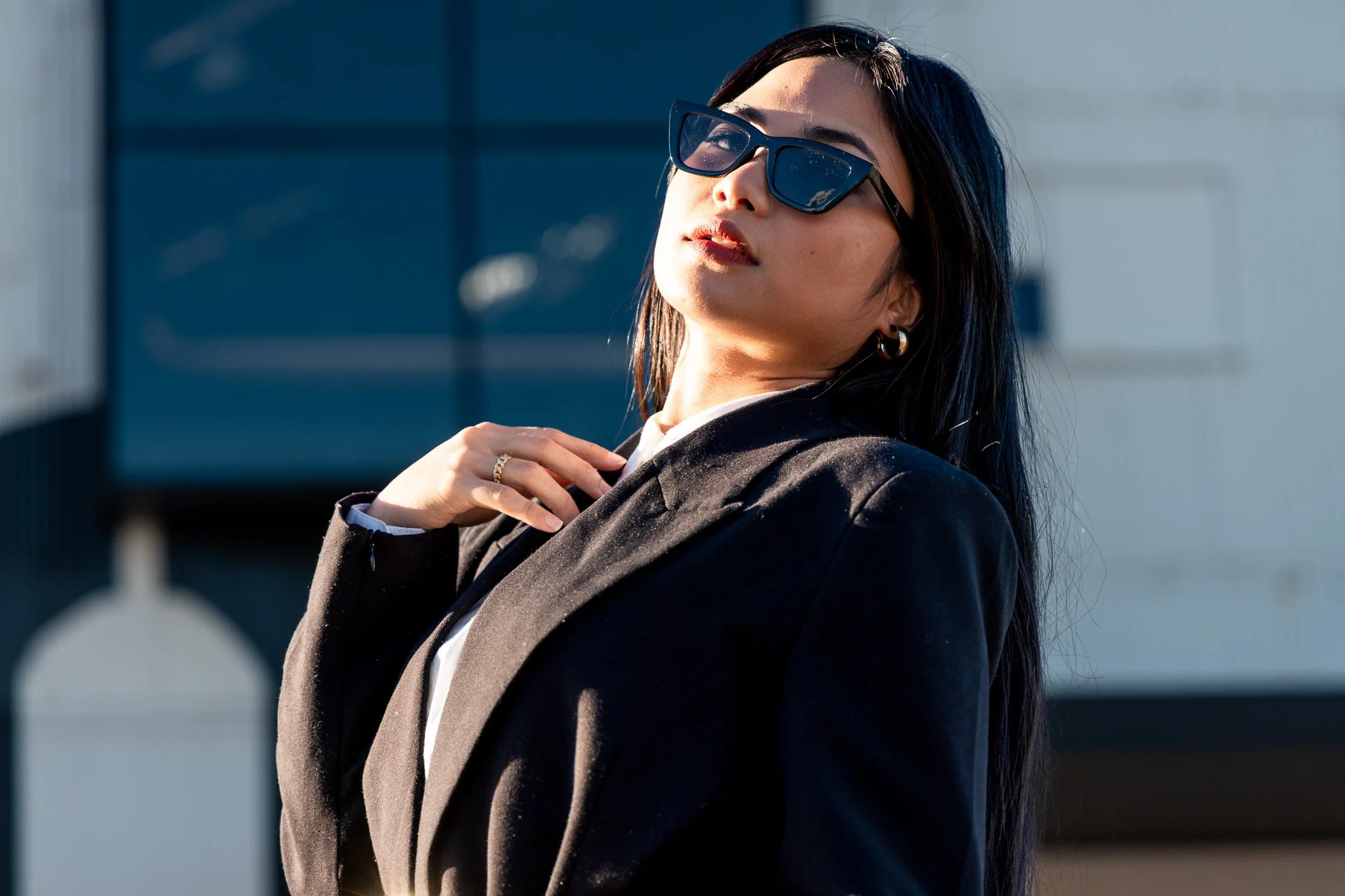 A woman with dark hair wearing large sunglasses, gold hoop earrings, and a black blazer over a white shirt, standing outdoors with a modern building in the background.