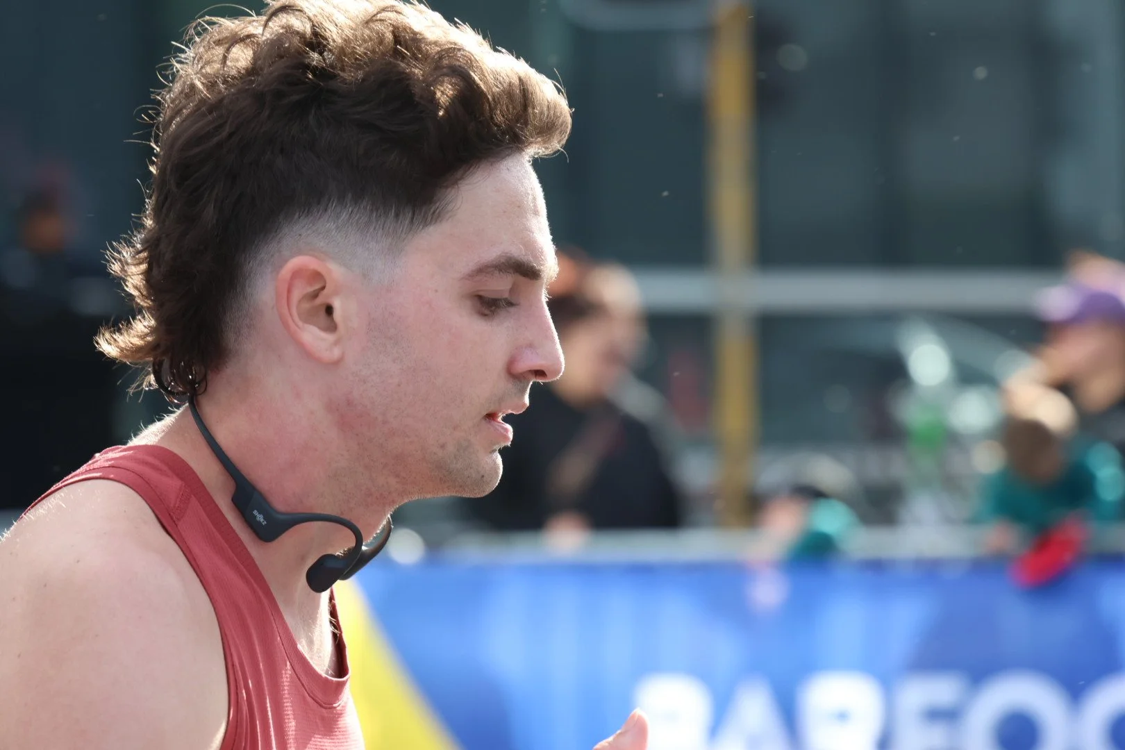 A young man with brown hair and a short beard wearing a red athletic tank top and a black neckband with a microphone at an outdoor running event, with a blurred background of spectators and structures.