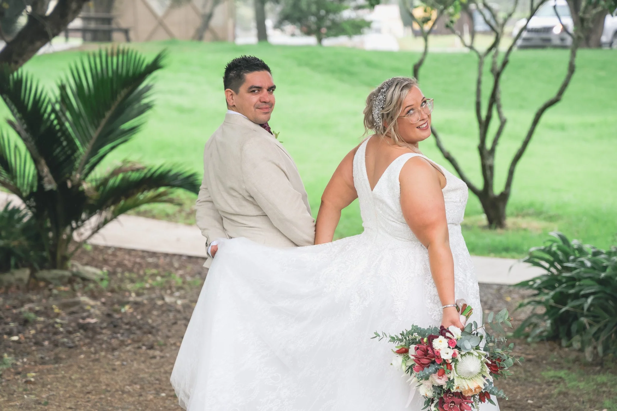A bride and groom sit on a bench outdoors in a park-like setting, with trees and grass in the background. The bride wears a white wedding dress and glasses, holds a bouquet of flowers, and turns her head to look back at the camera with a smile. The g