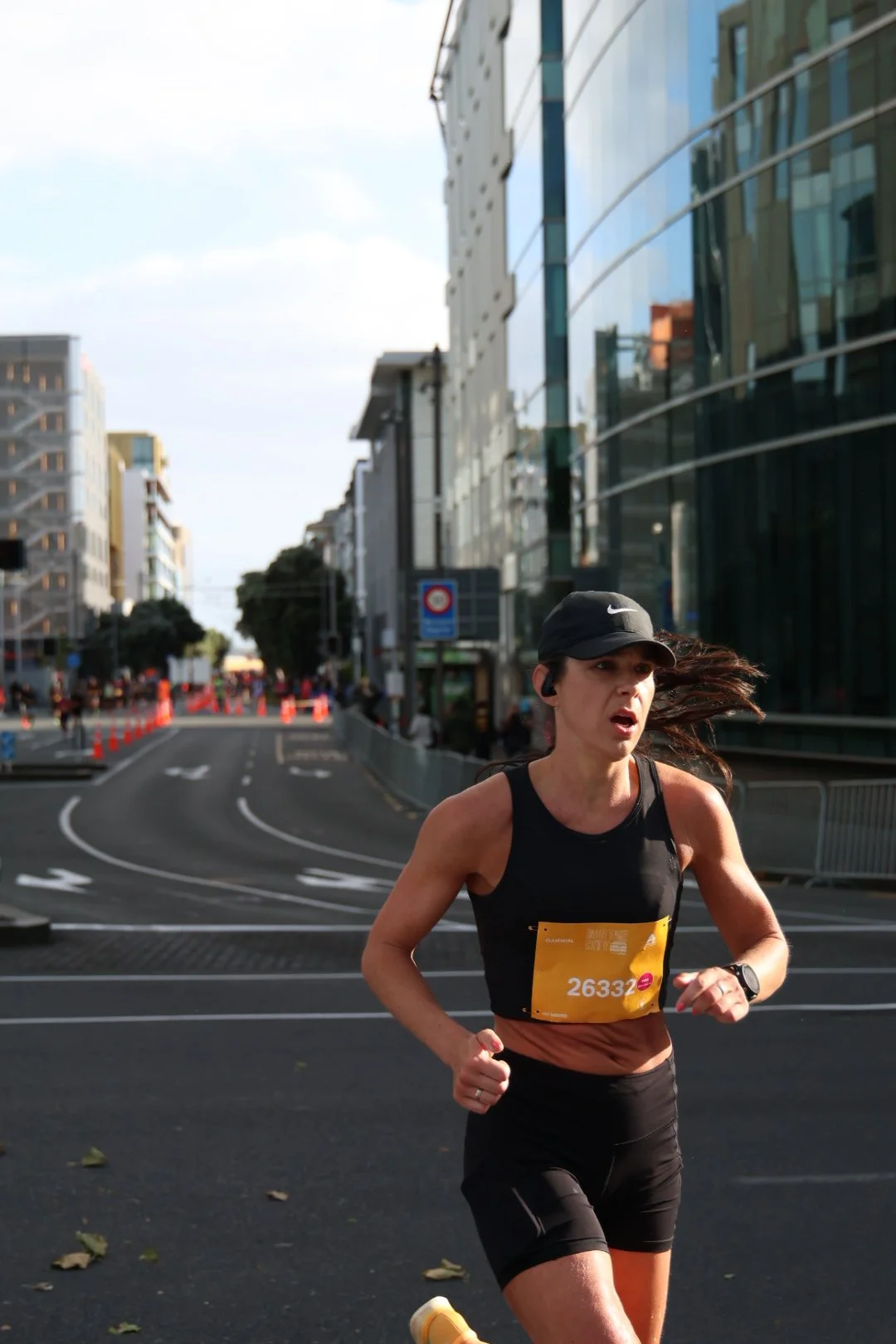 A woman running in a city street during a marathon, wearing a black athletic outfit, a black cap, and a yellow bib with the number 26332, with modern buildings and street signs in the background.