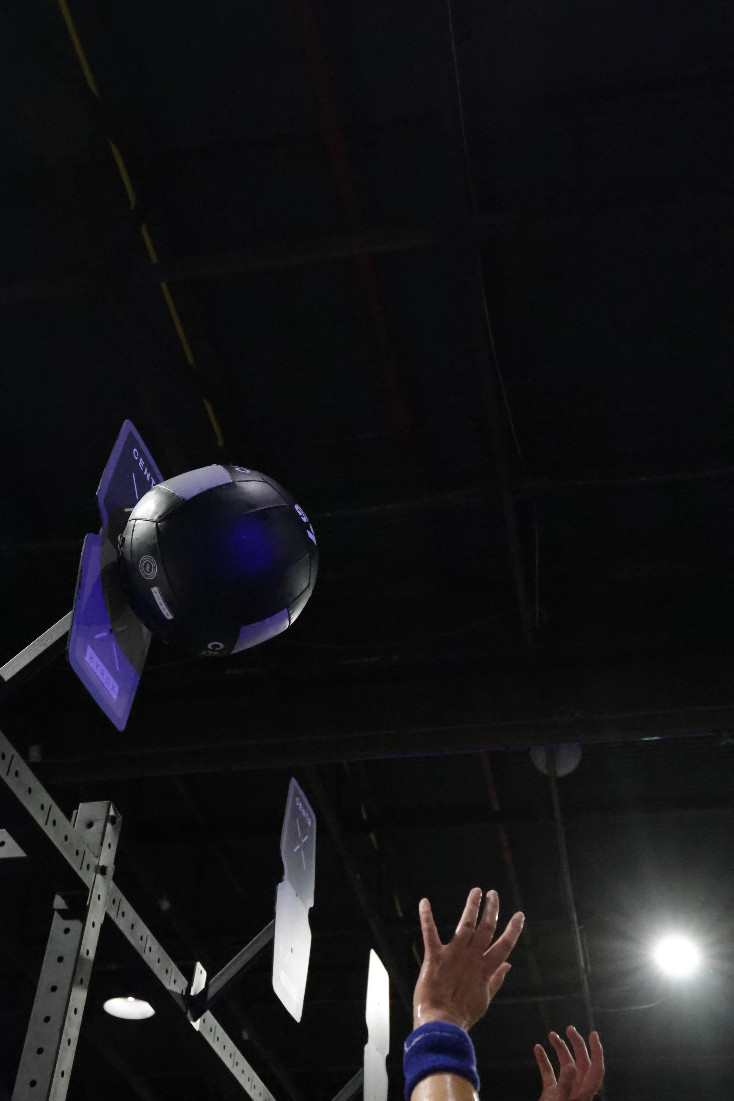 A dark indoor space with what appears to be fitness or gym equipment and a person's hand reaching up toward a hanging or floating object, possibly a helmet or a ball, with some signs and structures visible in the background.